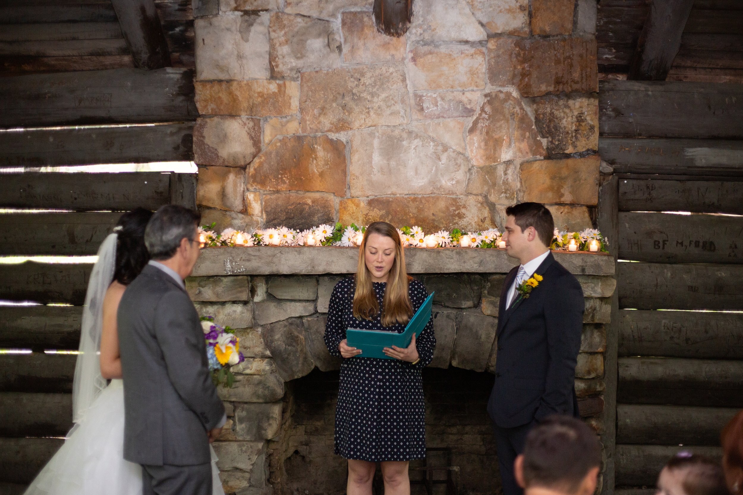 A wedding ceremony with four people standing in front of a stone fireplace, two men and one woman with a woman officiant reading from a book. The bride is in a white wedding dress, and the groom is in a dark suit with a sunflower boutonniere. The oth