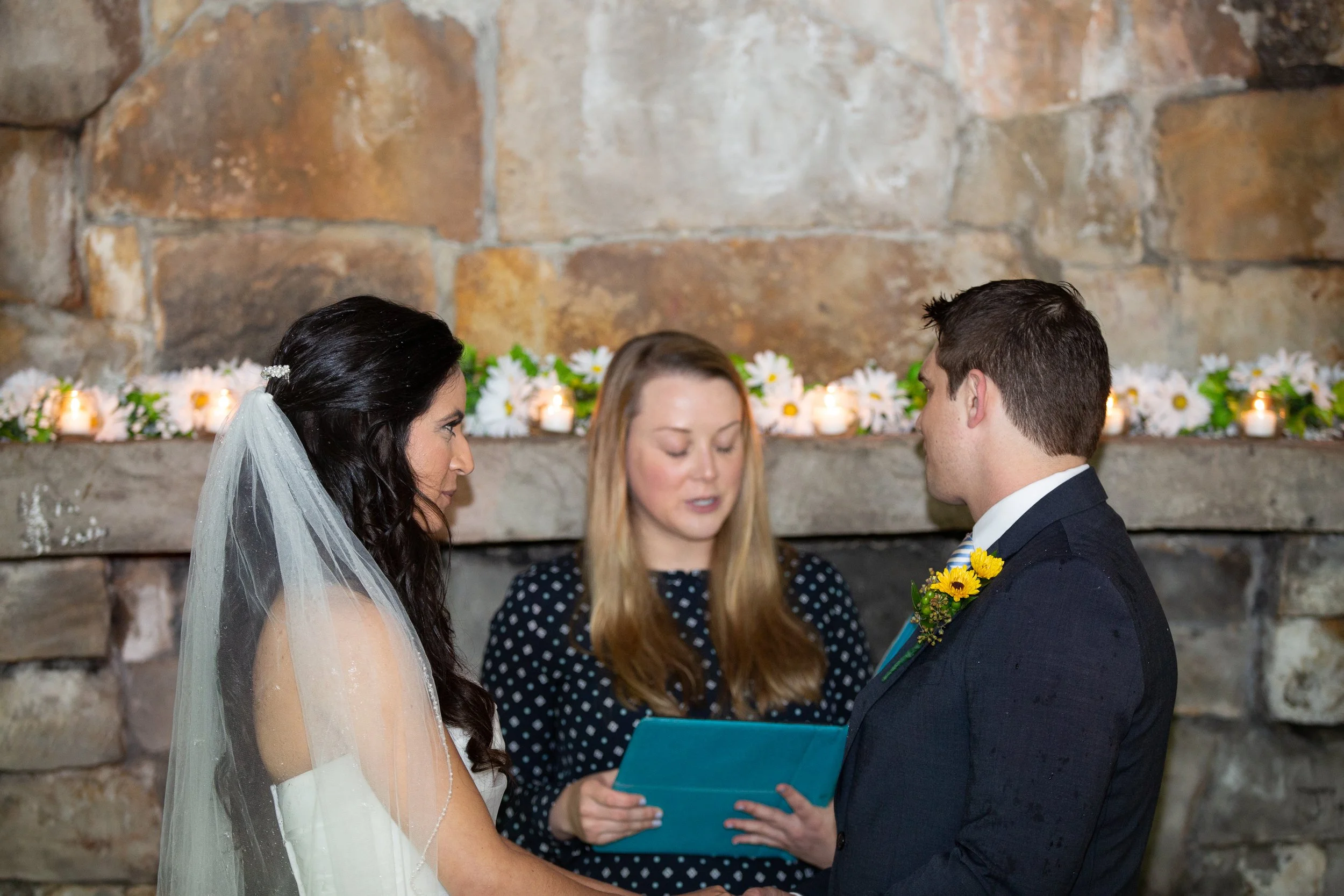 A bride and groom standing face to face during their wedding ceremony, with a woman officiating and a decorative background of candles and flowers.