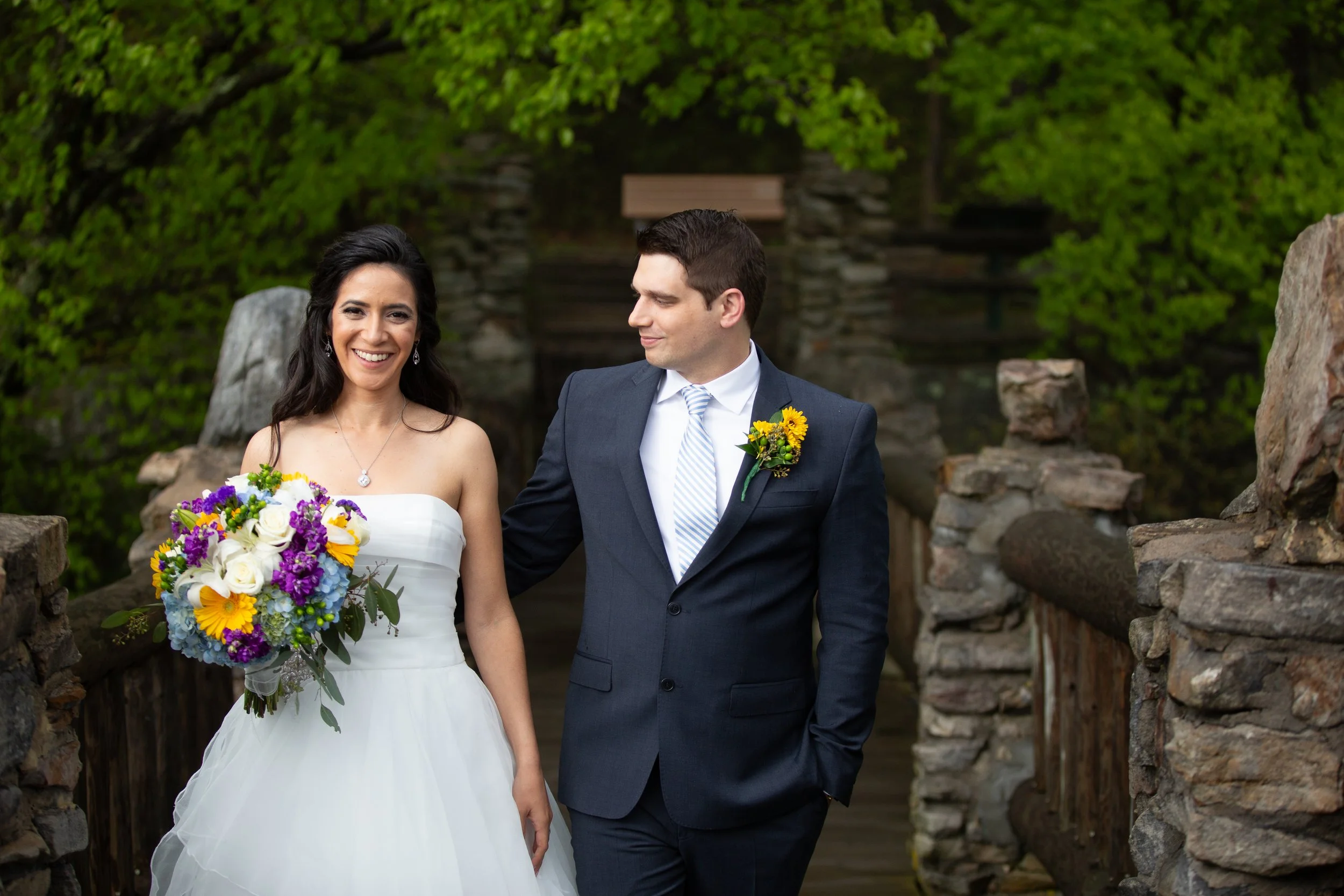 A bride and groom standing together outdoors near a stone wall, smiling, with the bride holding a colorful bouquet of flowers, surrounded by green trees.