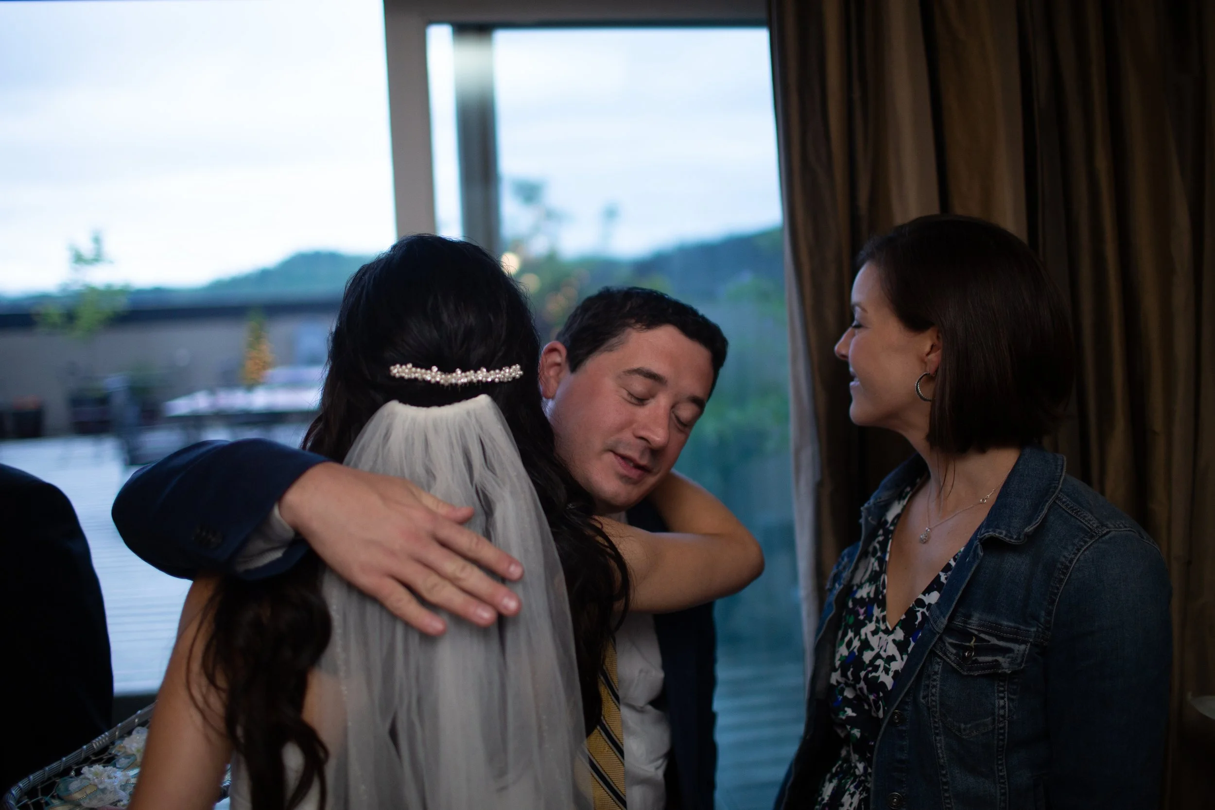 People sharing an emotional hug at a gathering, with a window and mountains in the background.