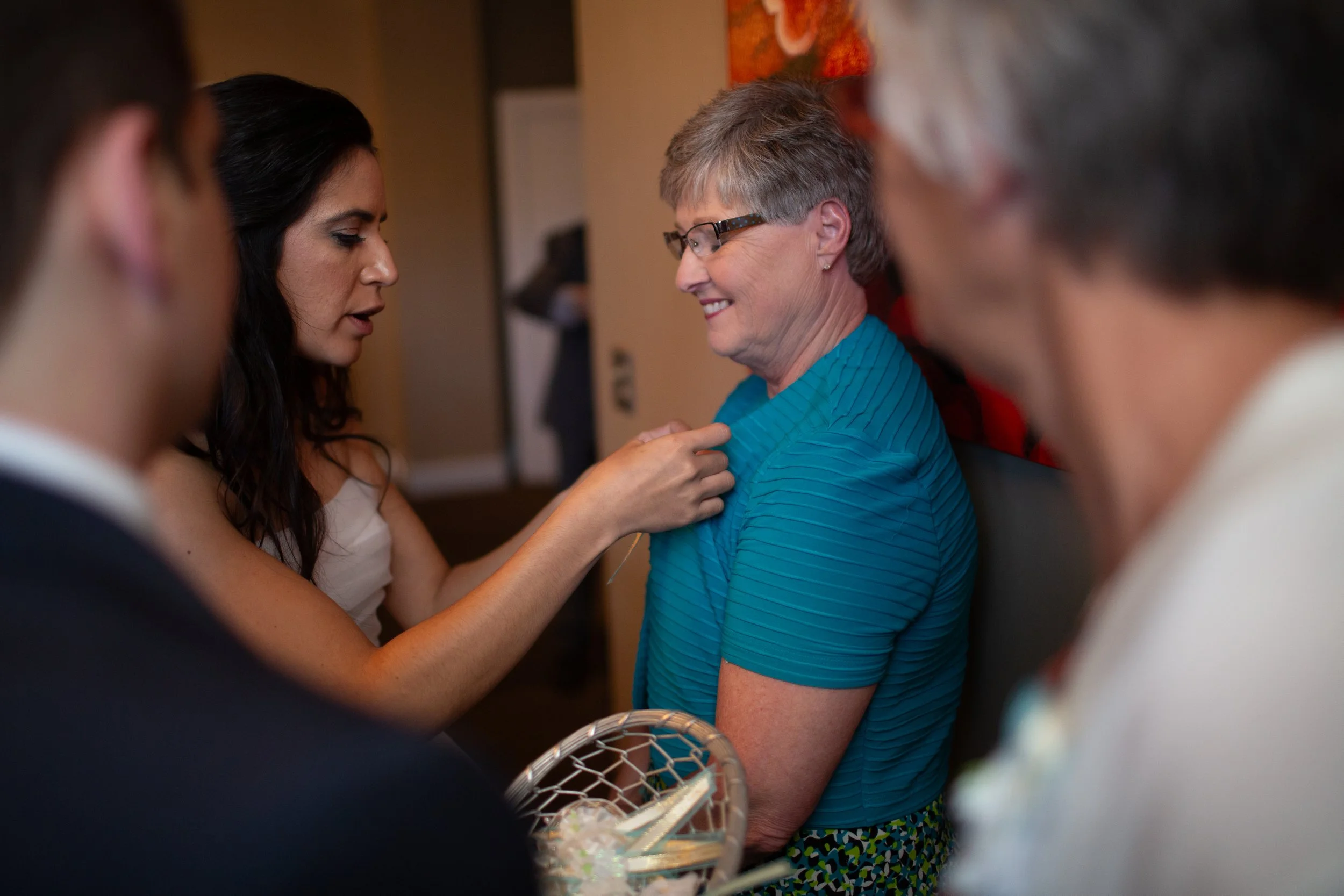 A woman in white dress pins a badge onto a smiling woman in glasses and a blue shirt, surrounded by blurred people.