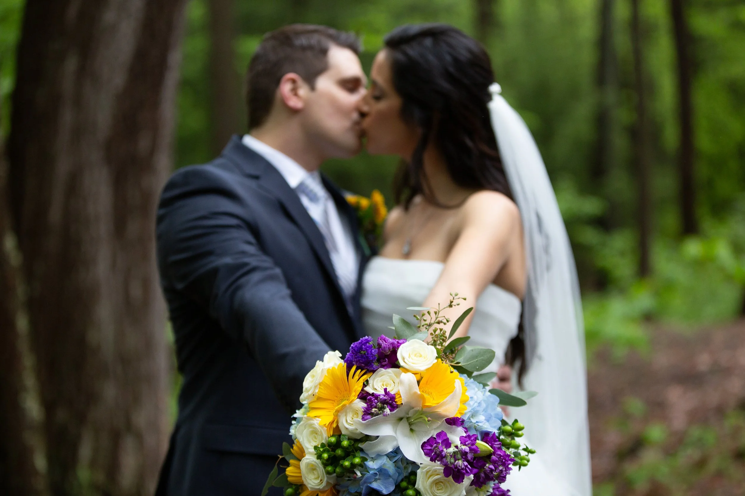 A bride and groom kissing during their wedding ceremony in a wooded outdoor setting, with the bride holding a colorful bouquet of flowers.
