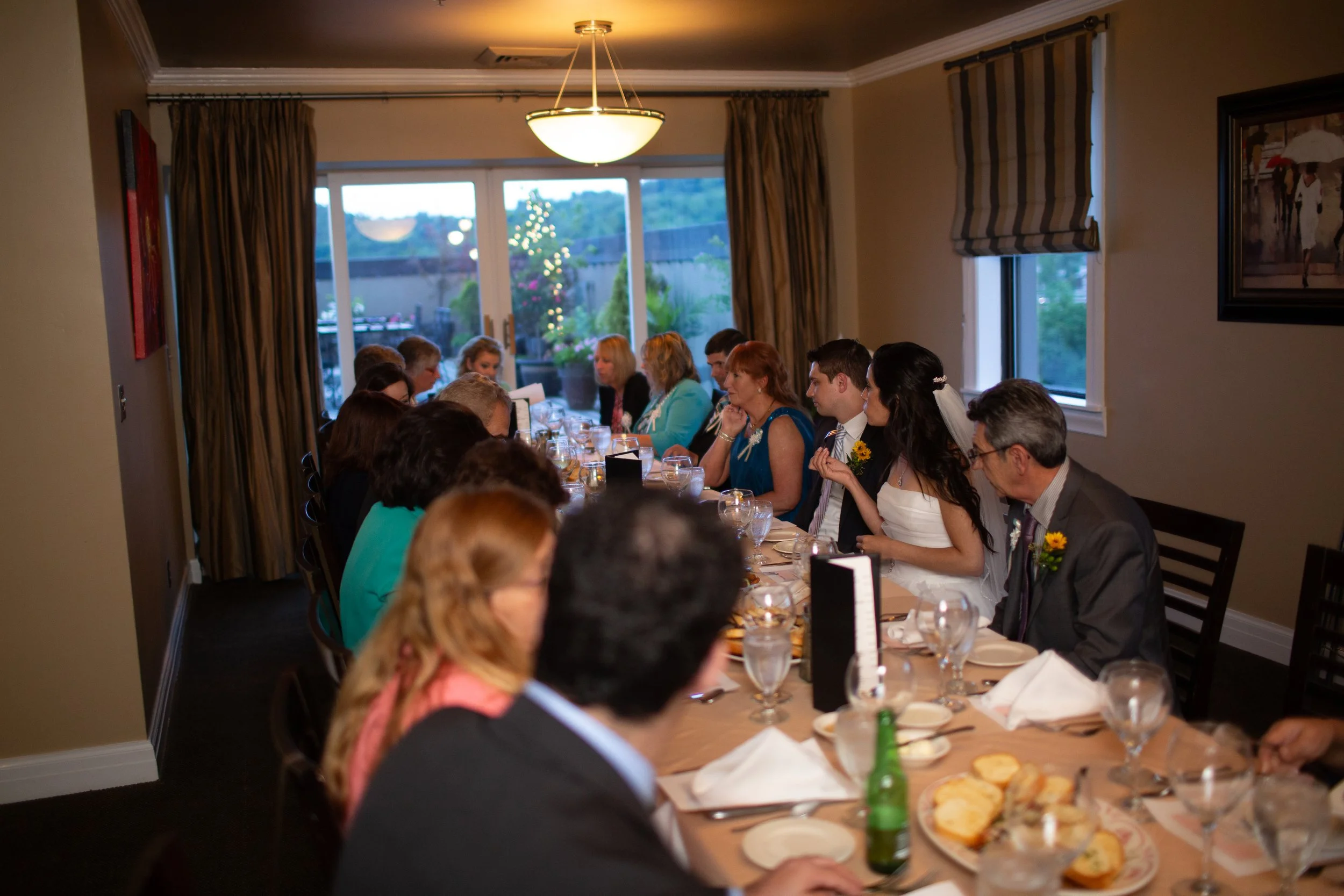 Guests sitting at a long dining table with white tablecloths, glasses, plates, and food in a well-lit dining room with windows showing an outdoor patio and a decorated Christmas tree.