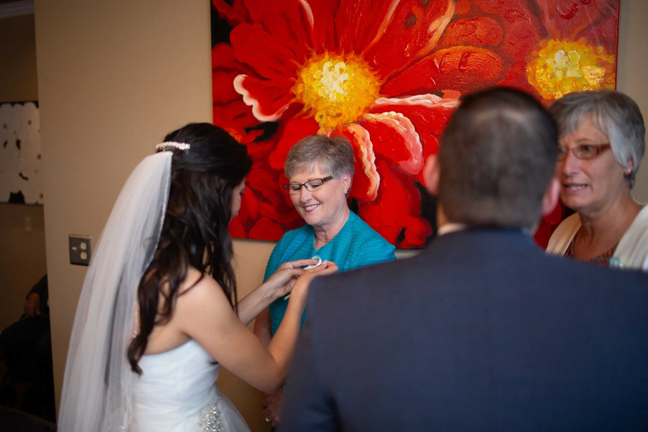 A bride and groom exchanging rings at a wedding reception with guests, including an older woman in a teal dress, in front of a large red and yellow flower painting on the wall.