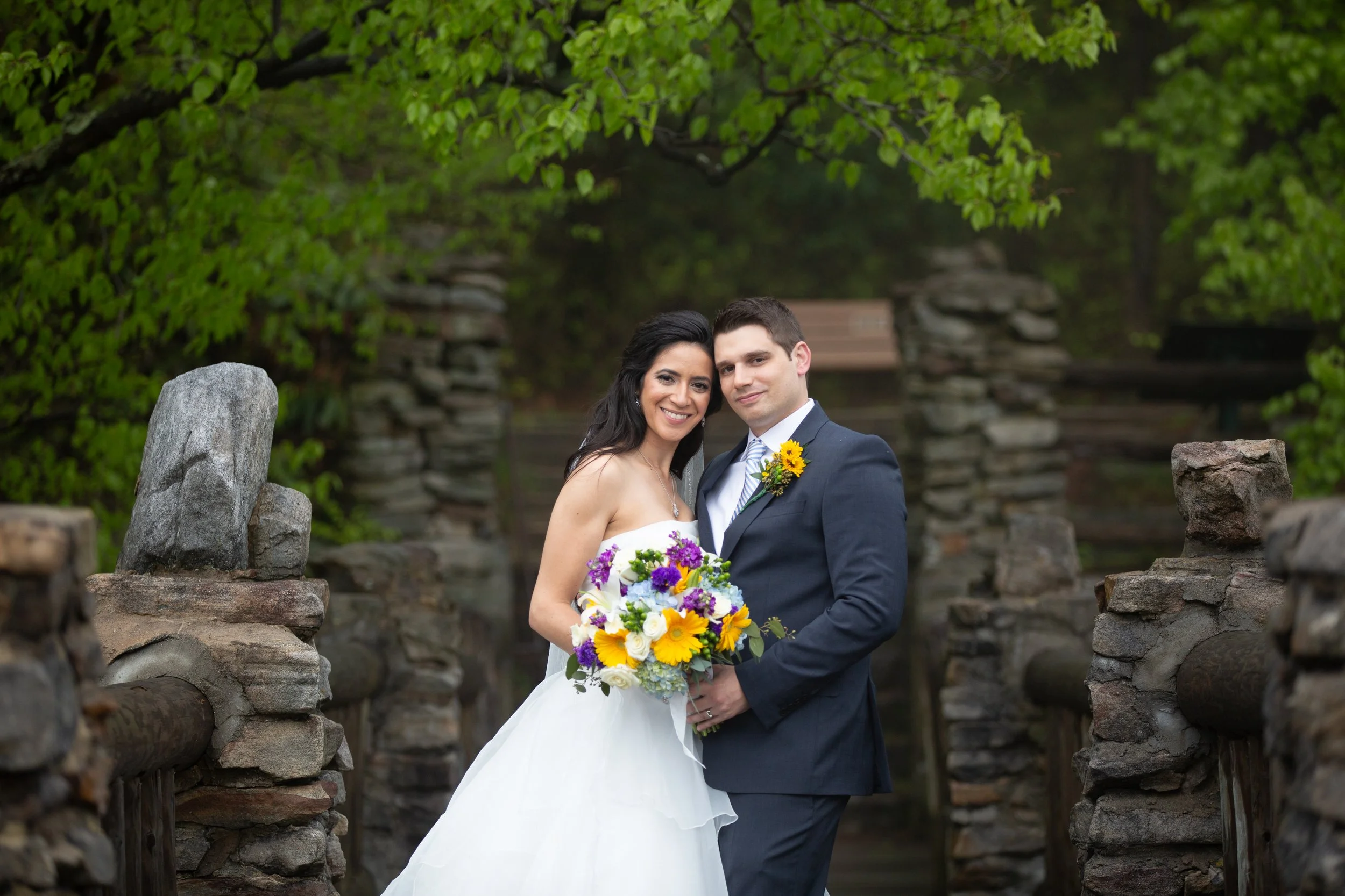 A bride and groom stand close together, smiling, outdoors among stone and greenery, with the bride holding a colorful bouquet of flowers.