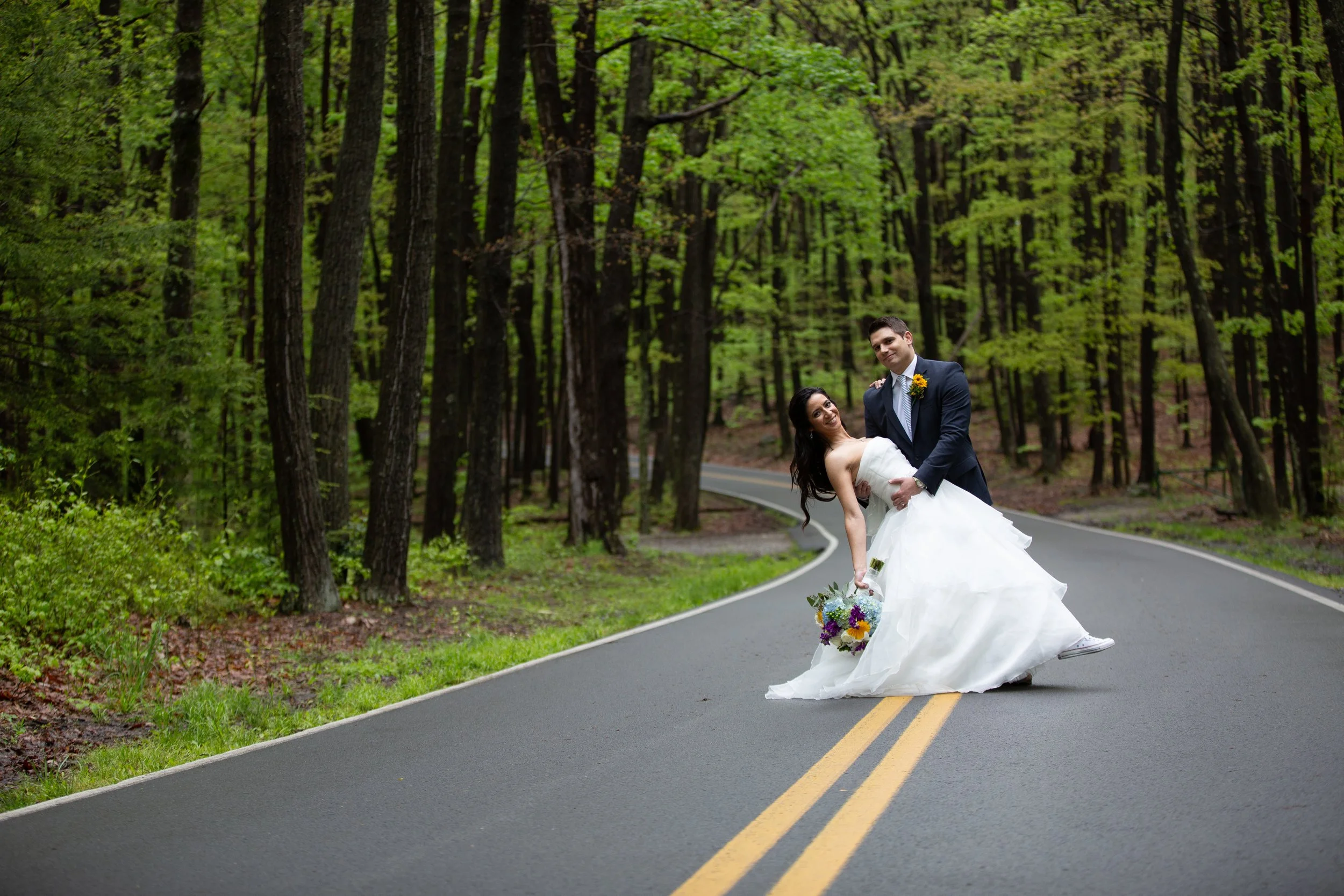 A newlywed couple in wedding attire dancing on a winding country road surrounded by a green forest.