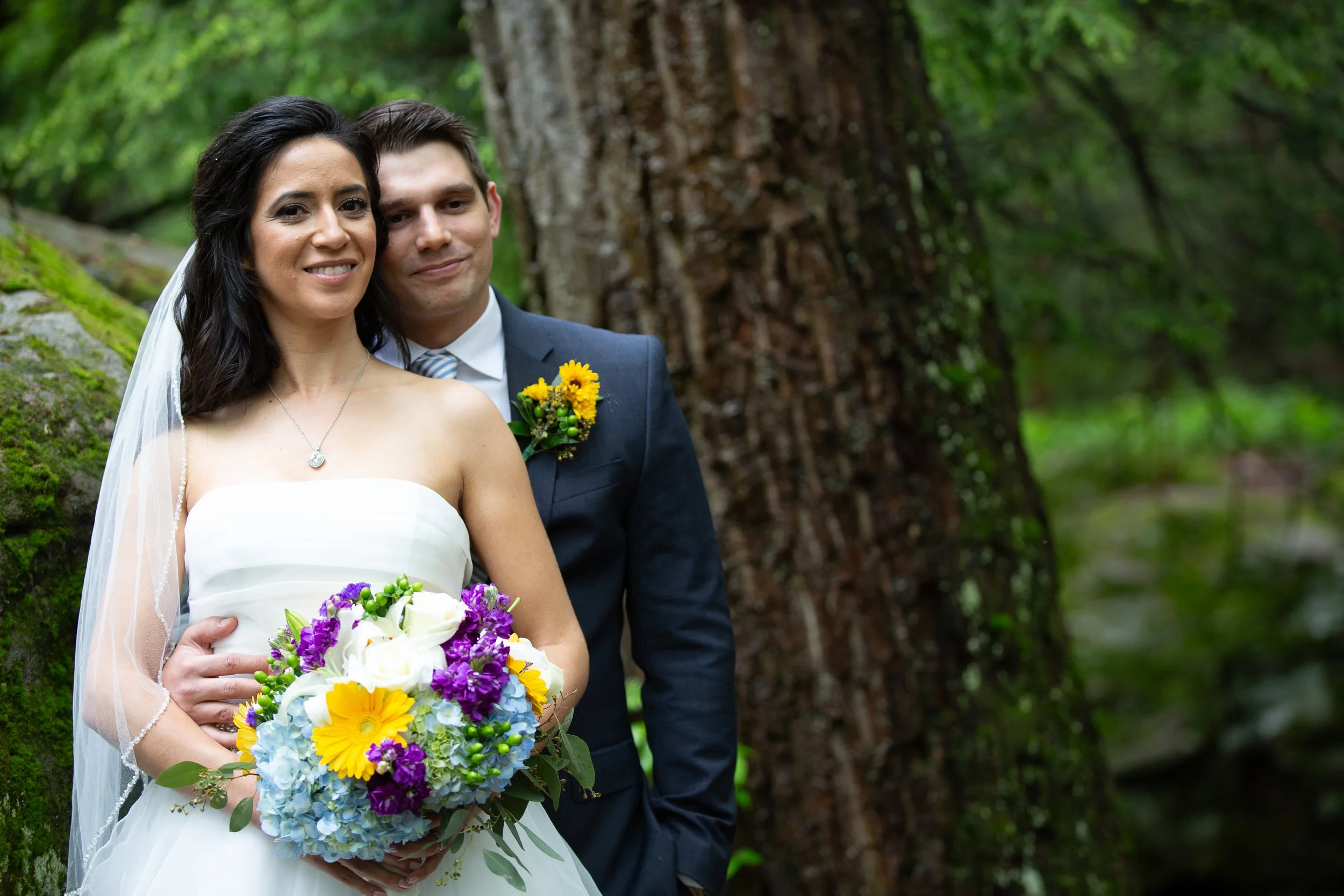 A bride and groom standing outdoors in a wooded area, smiling at the camera. The bride is holding a colorful bouquet of flowers, and the groom is wearing a dark suit with a striped tie and a yellow boutonniere. The background features green foliage a