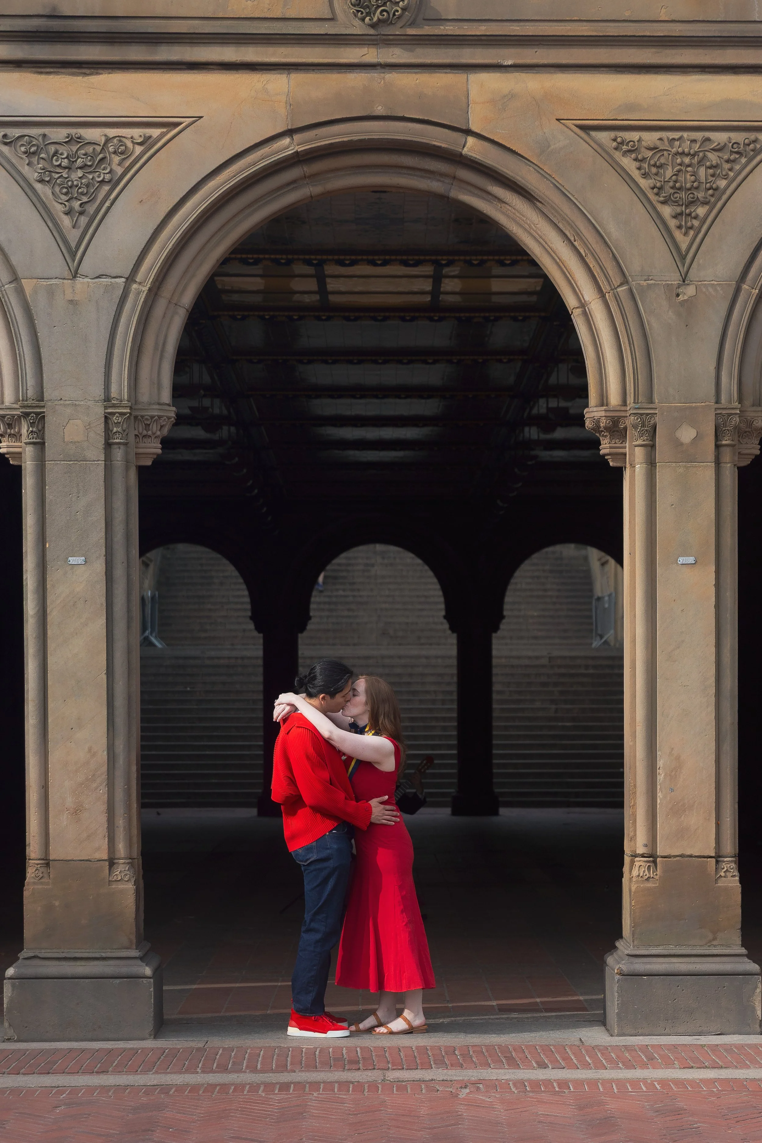 A couple is standing close together in front of a historic archway. They are touching foreheads, with the man wearing a red sweater and jeans, the woman in a long red dress and sandals. The background features stone columns, arches, and stairs.