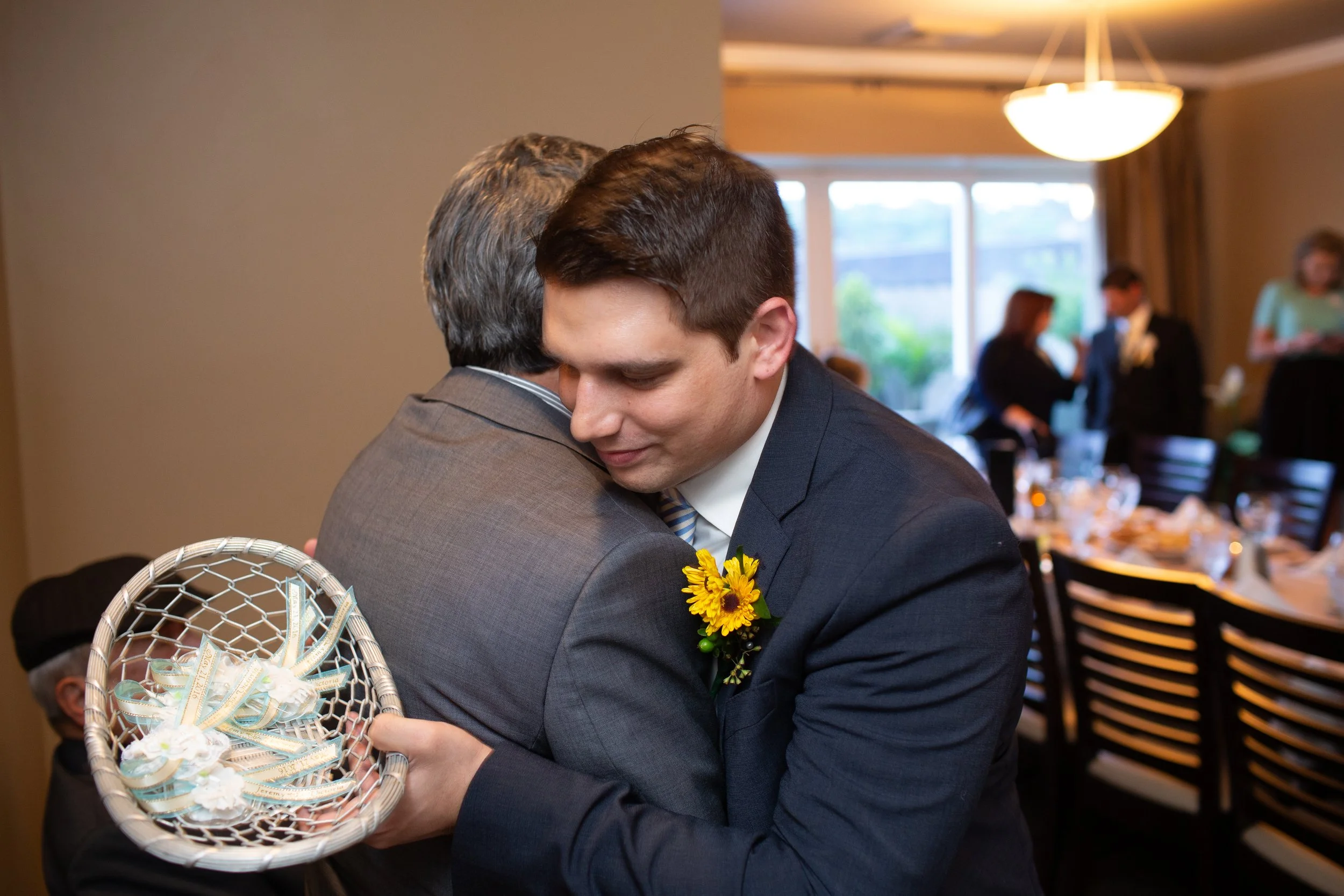 A young man in a suit with a sunflower boutonniere is hugging an older man. The young man is holding a basket of decorated white flowers and ribbons. In the background, there are people standing and sitting in a well-lit room.