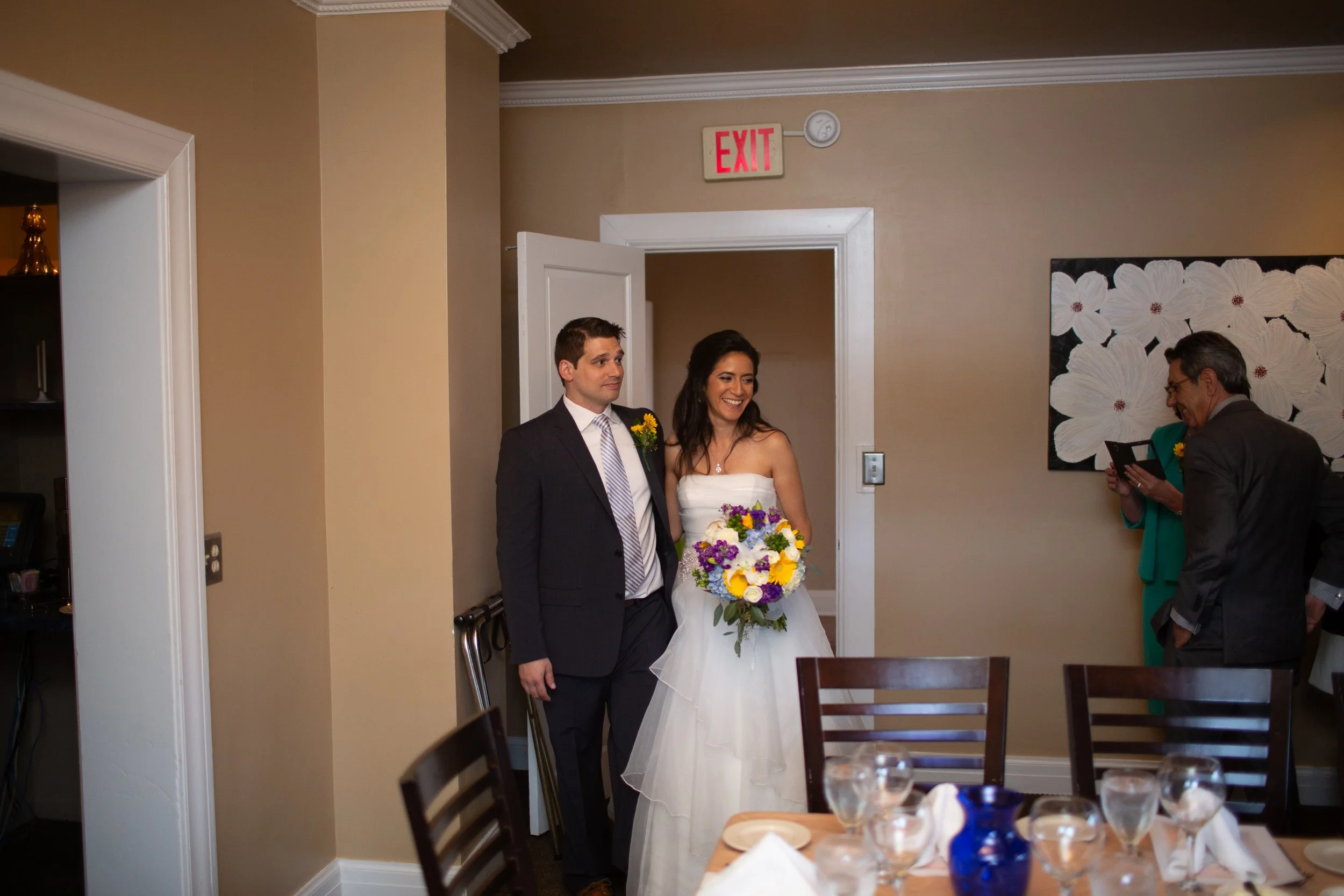 A bride and groom standing together, with the bride holding a bouquet of colorful flowers, at a wedding reception. A woman is seen smiling and looking at them, and a man wearing glasses is also present.