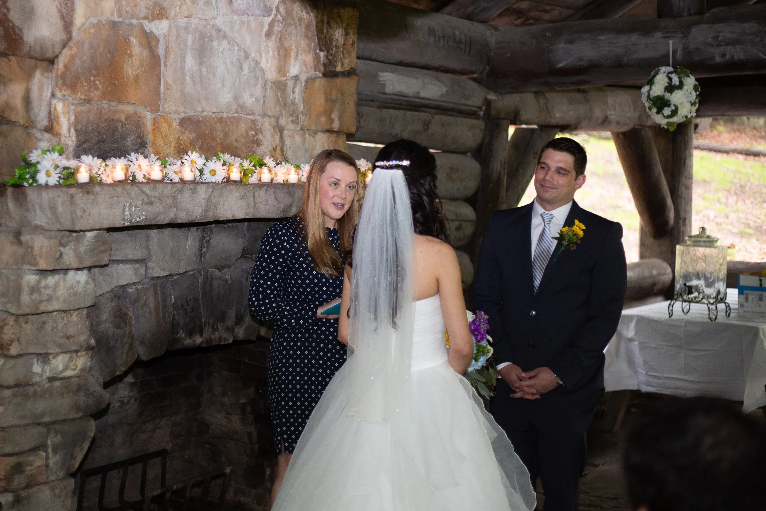 A wedding ceremony with a bride and groom standing together with an officiant or friend. The bride is wearing a white wedding dress and veil, the groom is in a dark suit with a striped tie and yellow boutonniere. They are inside a rustic stone and lo