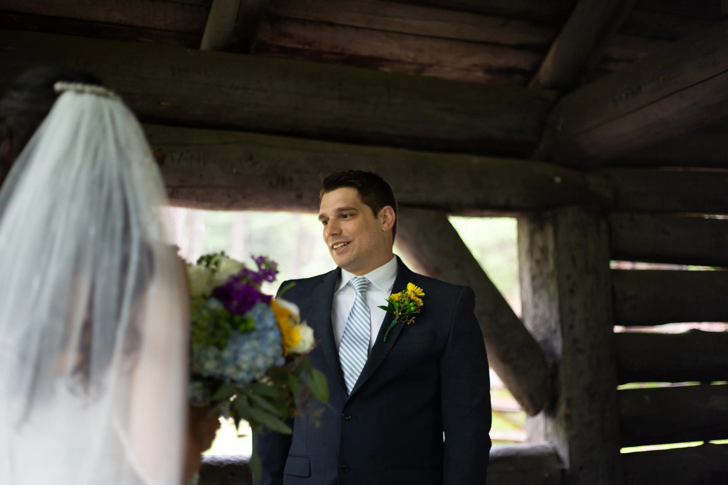 A man in a dark suit and striped tie smiling during a wedding ceremony, standing under a wooden structure with out-of-focus bride in the foreground holding a bouquet of flowers.