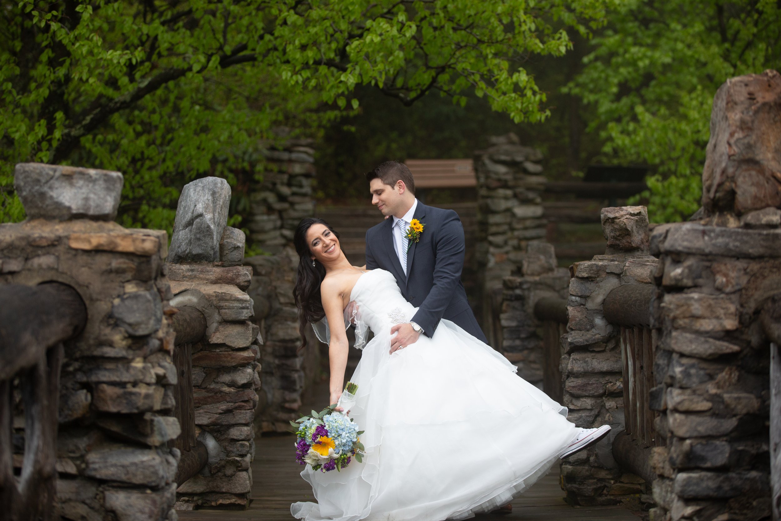 A bride in a white wedding gown is being held by a groom in a suit as they smile and look at each other near a stone bridge with lush green trees surrounding them.