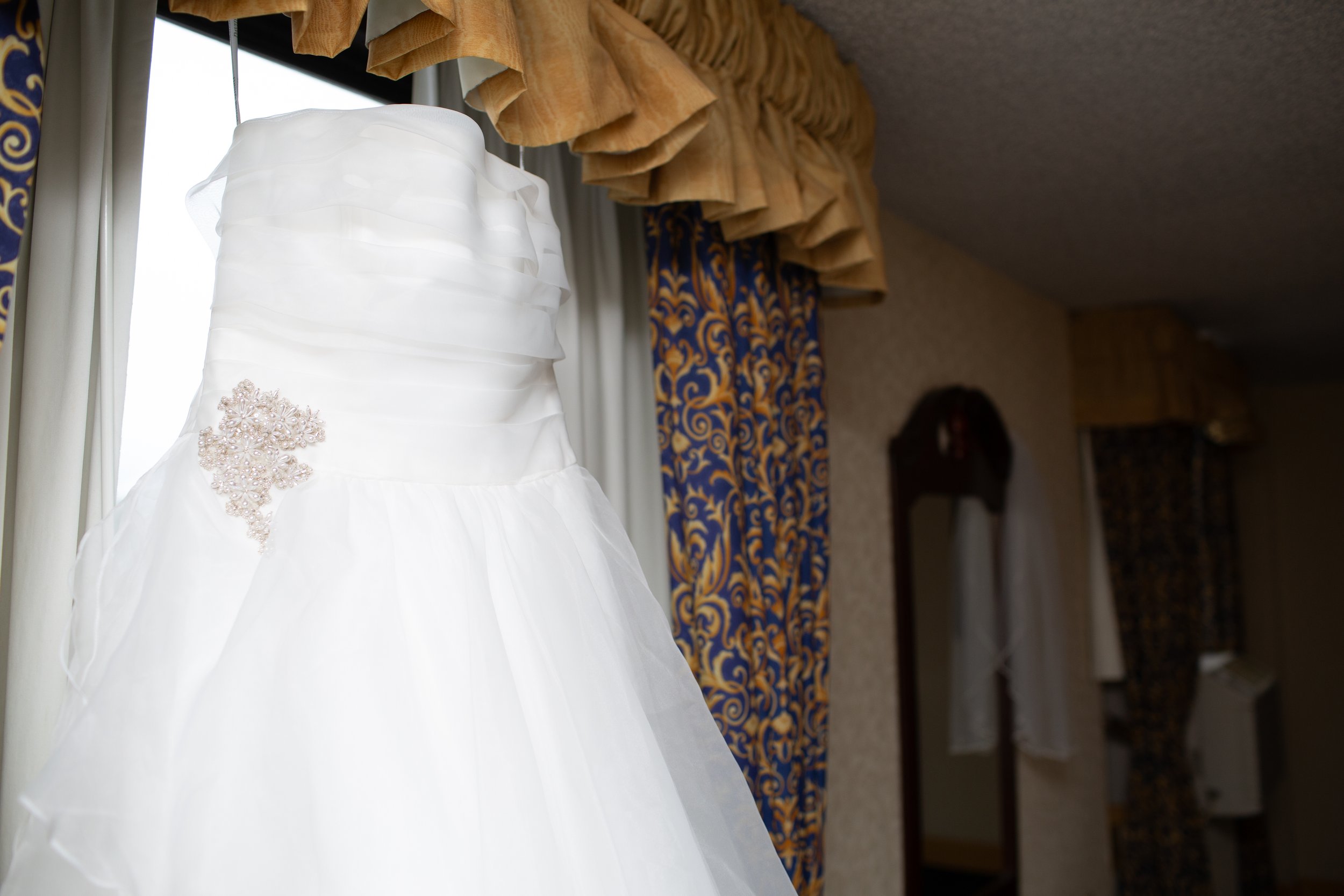 Close-up of a white wedding dress with lace embellishment, hanging near a window with patterned curtains and gold-colored valance, with a blurred background of a room.