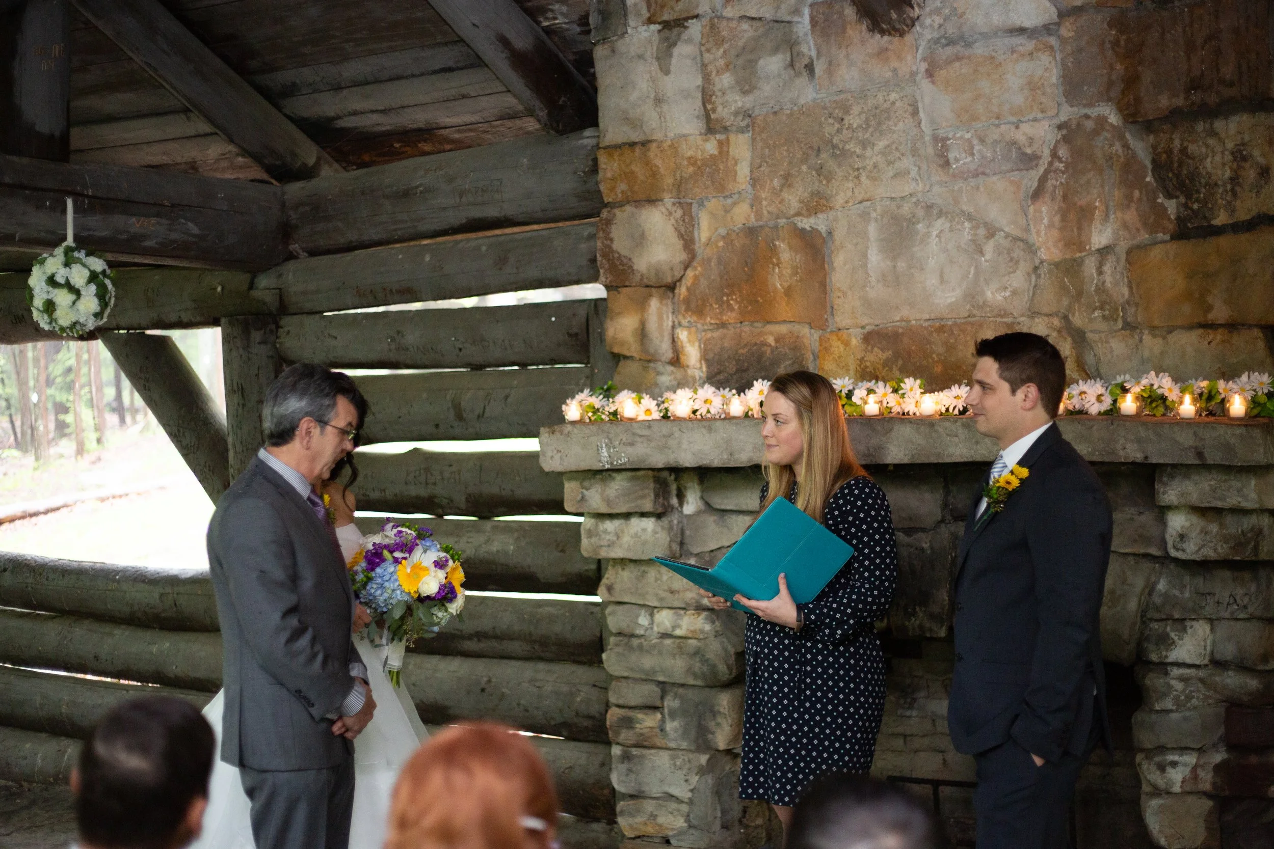 A wedding ceremony taking place inside a rustic log and stone building, with three people standing at the altar, one holding flowers, and others reading vows.