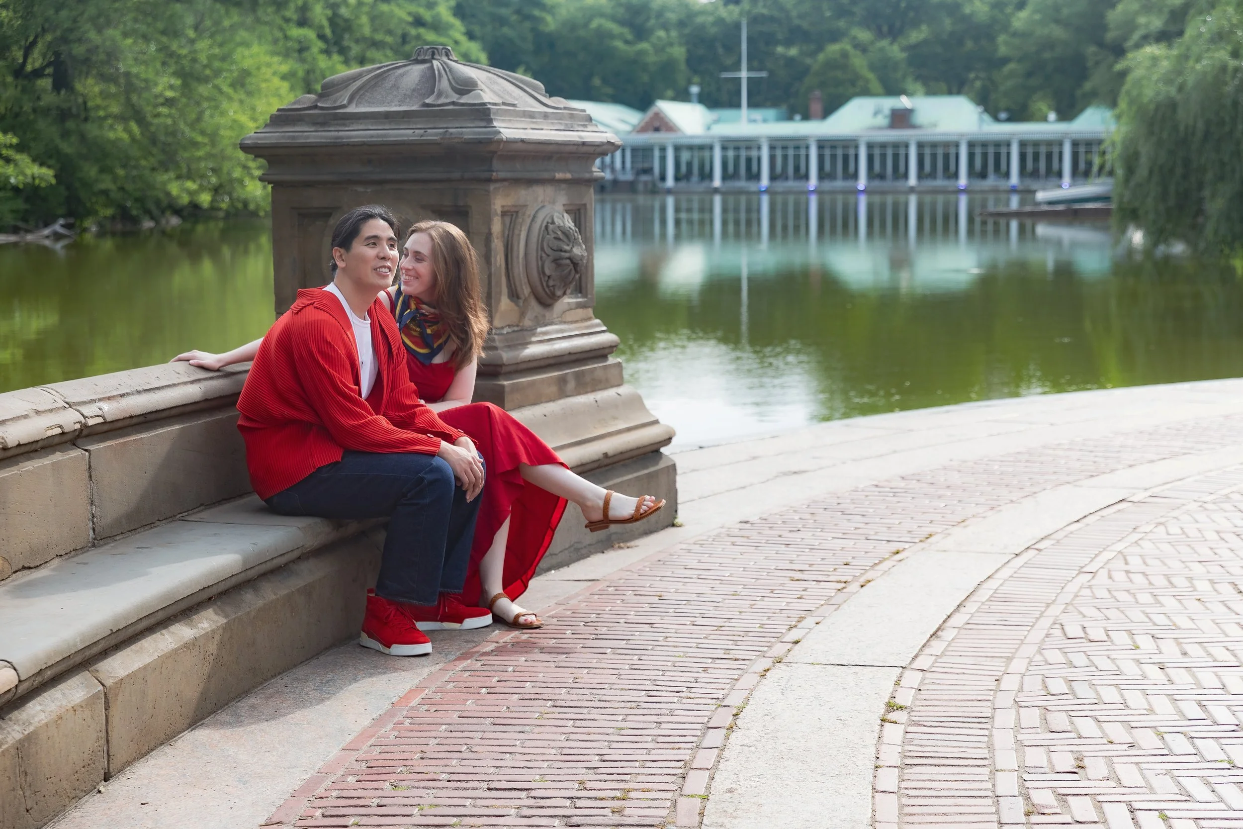 A young couple sitting on a stone railing by a river in a park, surrounded by green trees and a building in the background, smiling and enjoying each other's company.