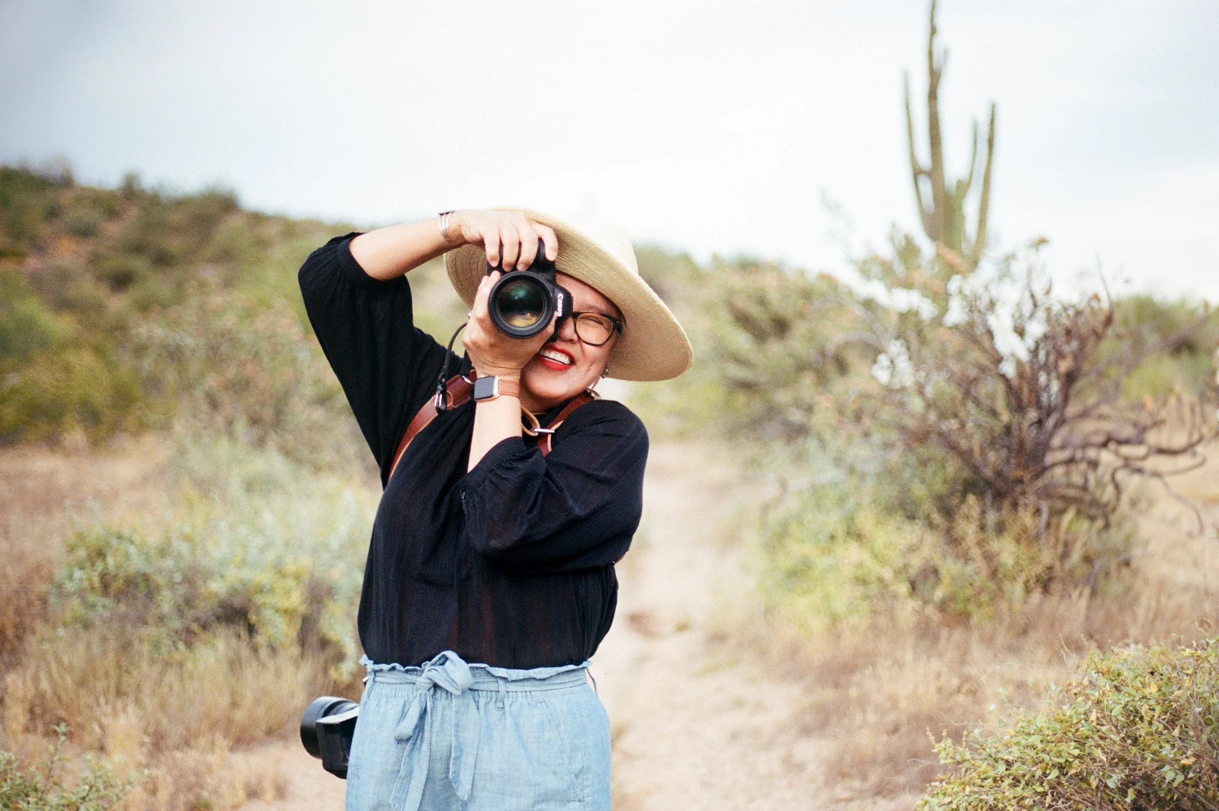 Woman in a wide-brimmed hat taking a photograph outdoors in a rural area with desert plants and a dry dirt path.