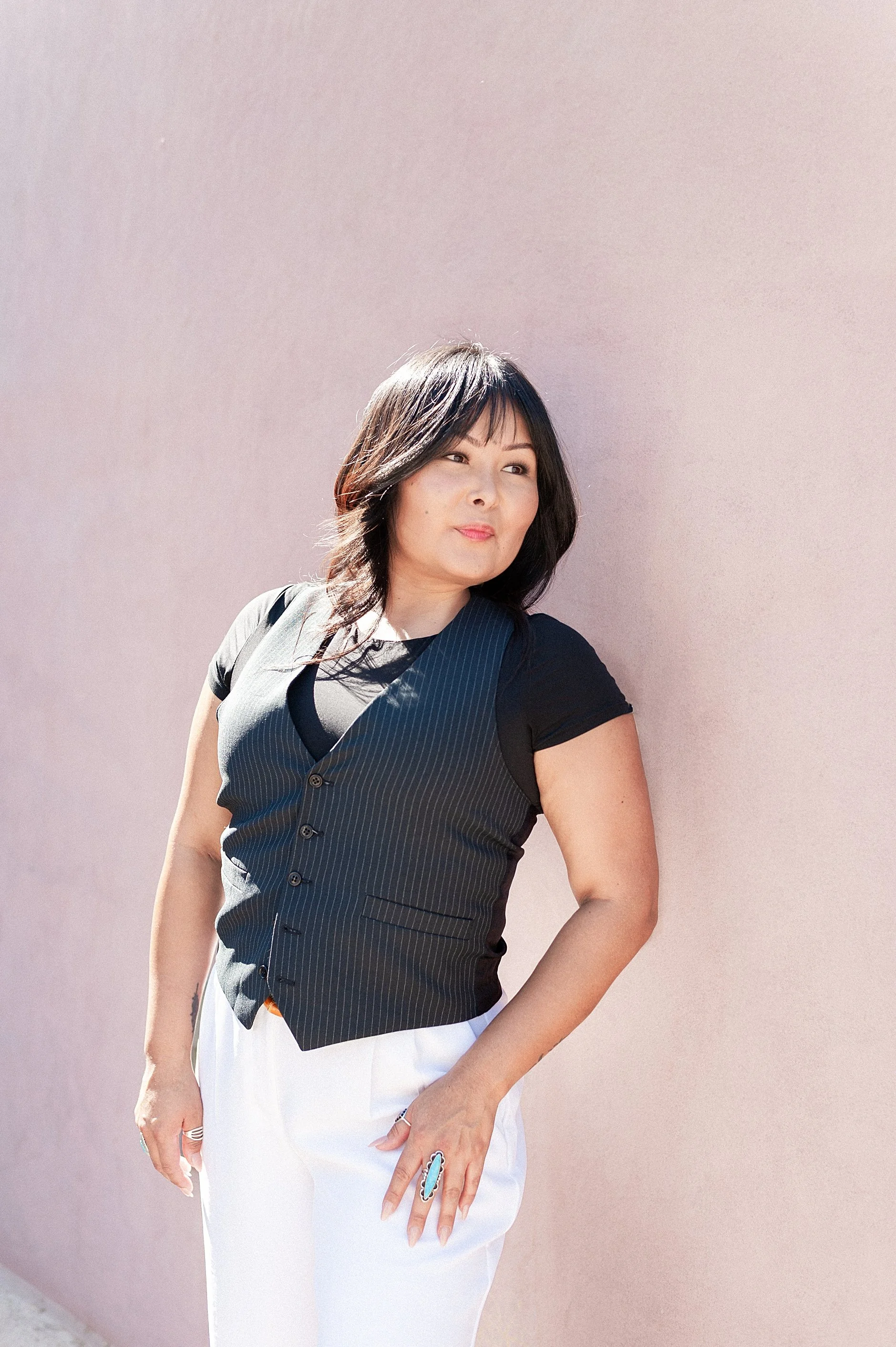 A woman with dark hair standing against a pale pink wall, wearing a dark striped vest and white pants.