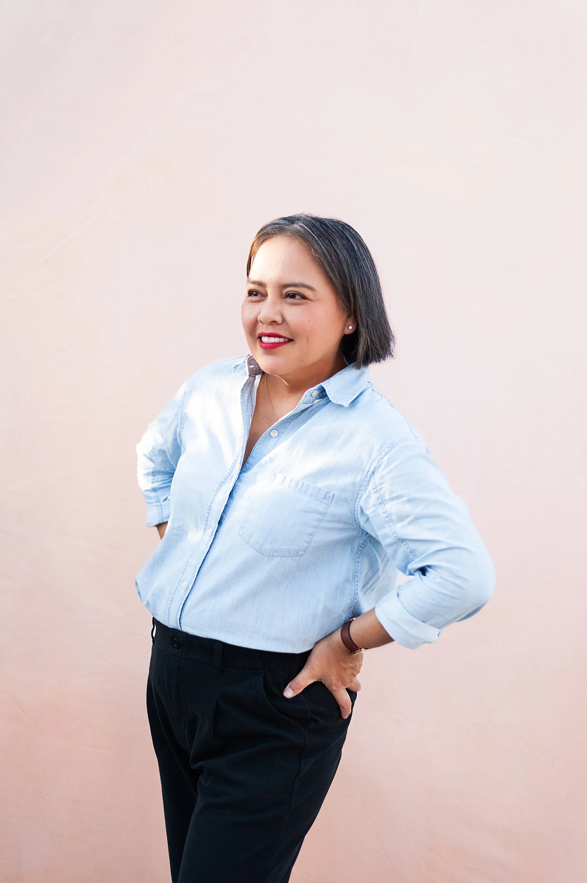 A woman with short dark hair, wearing a light blue button-up shirt and black pants, standing against a plain beige background. She has a confident expression, with her left hand on her hip and her right arm relaxed.