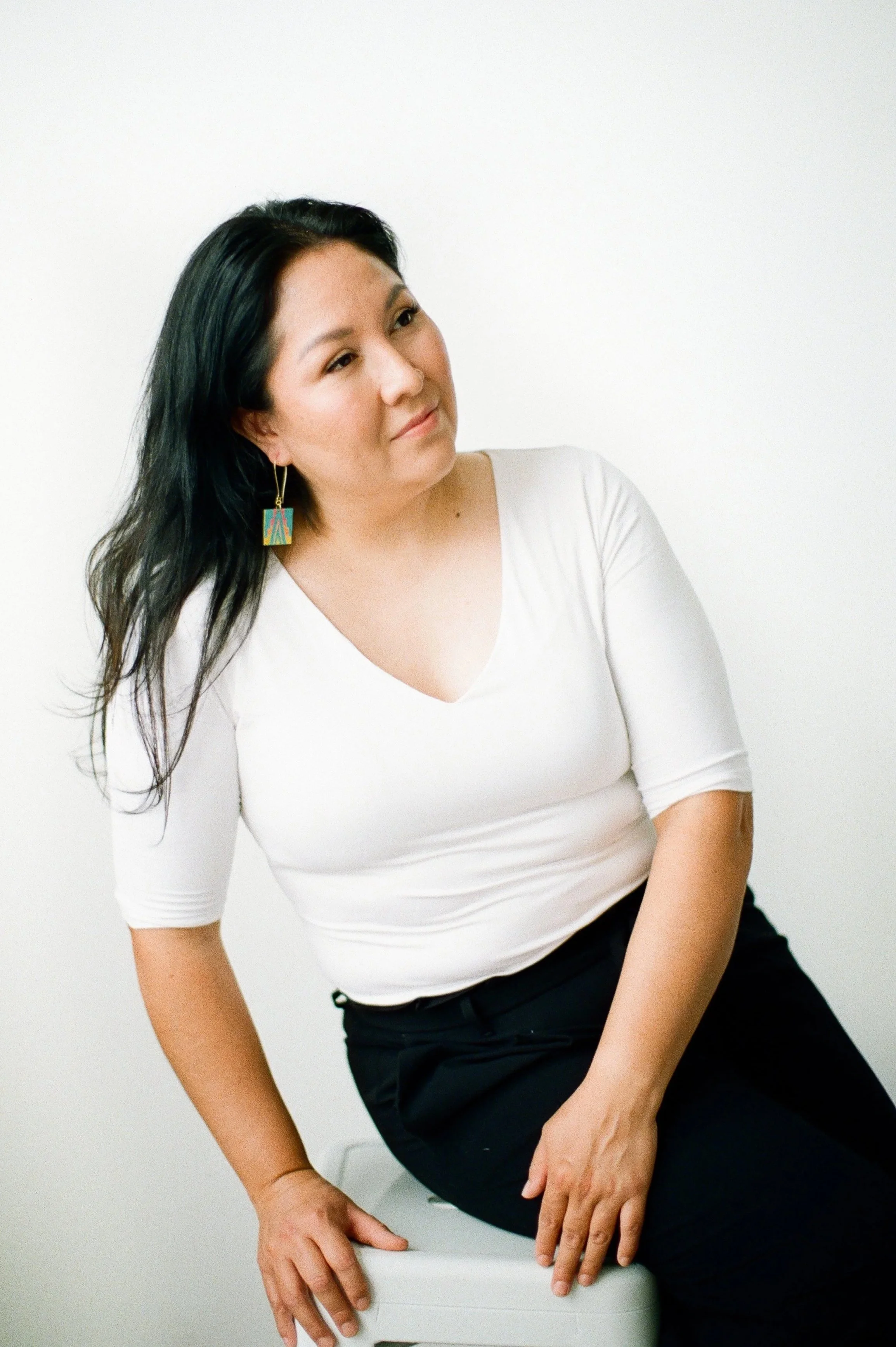 A woman with long dark hair sitting on a white stool, wearing a white top and black pants, against a plain white background.
