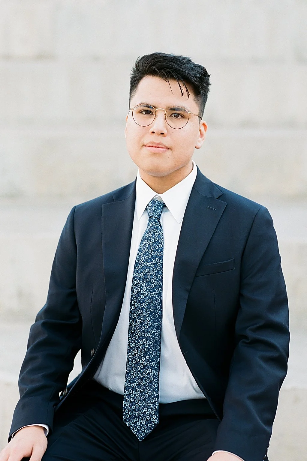 Young man wearing glasses in a dark suit, white shirt, and patterned tie, sitting against a neutral background.