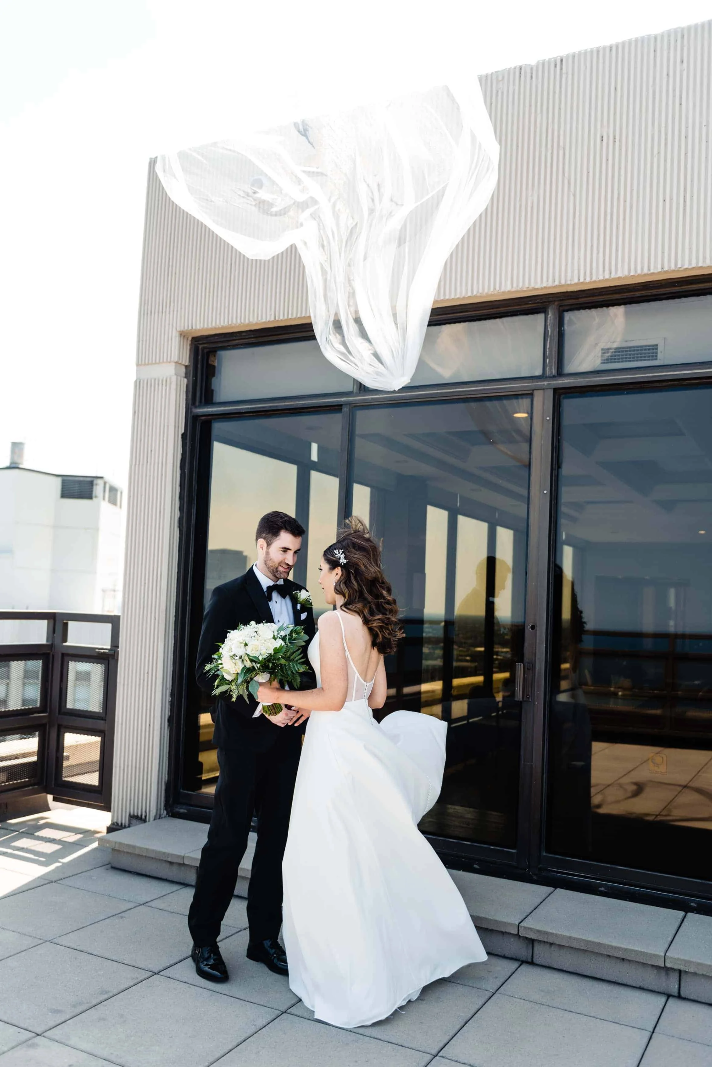 bride and groom stand on a rooftop and her veil blows away