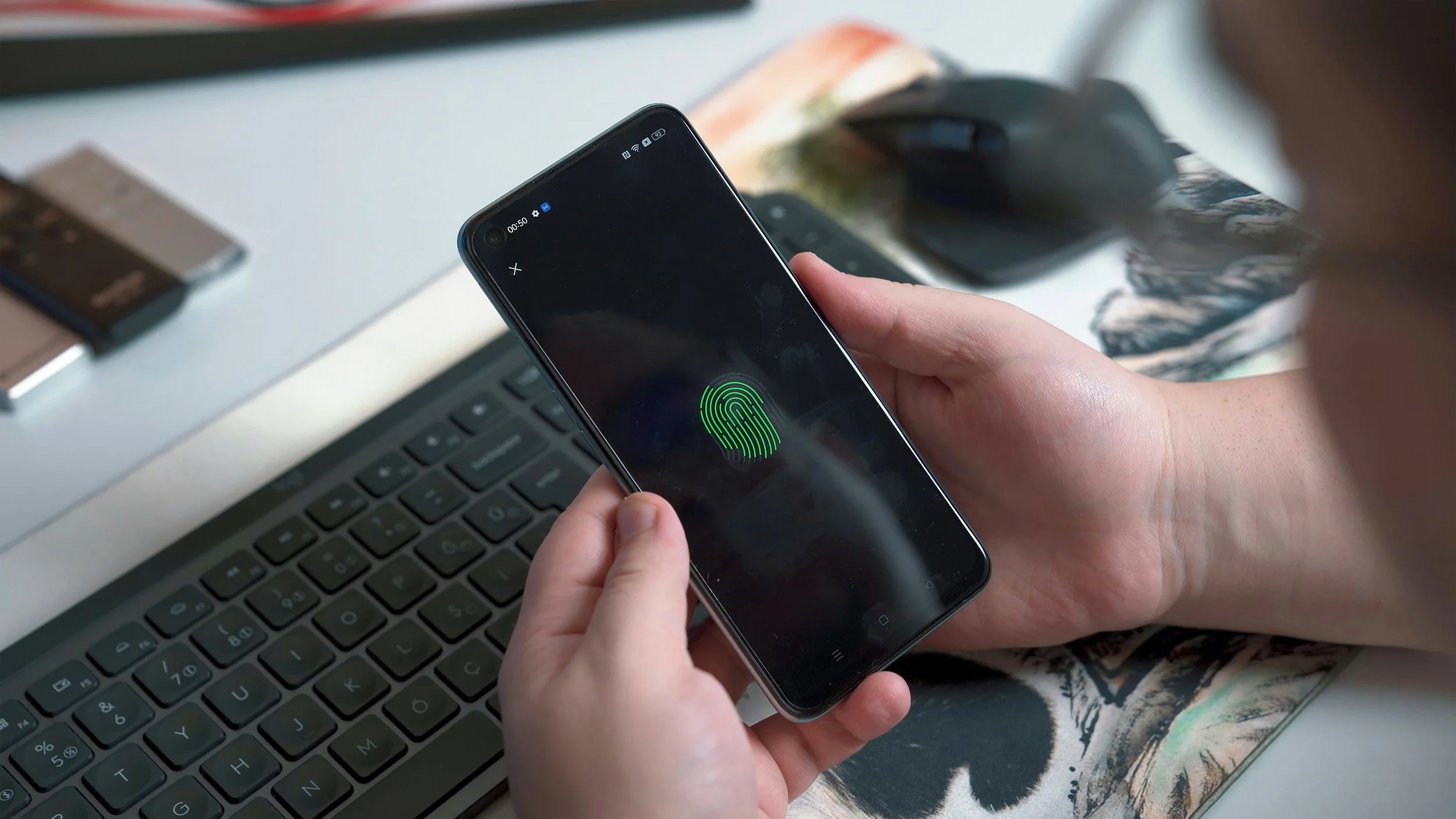 Person holding a smartphone displaying a biometric fingerprint scan, with a laptop and mouse on a desk.