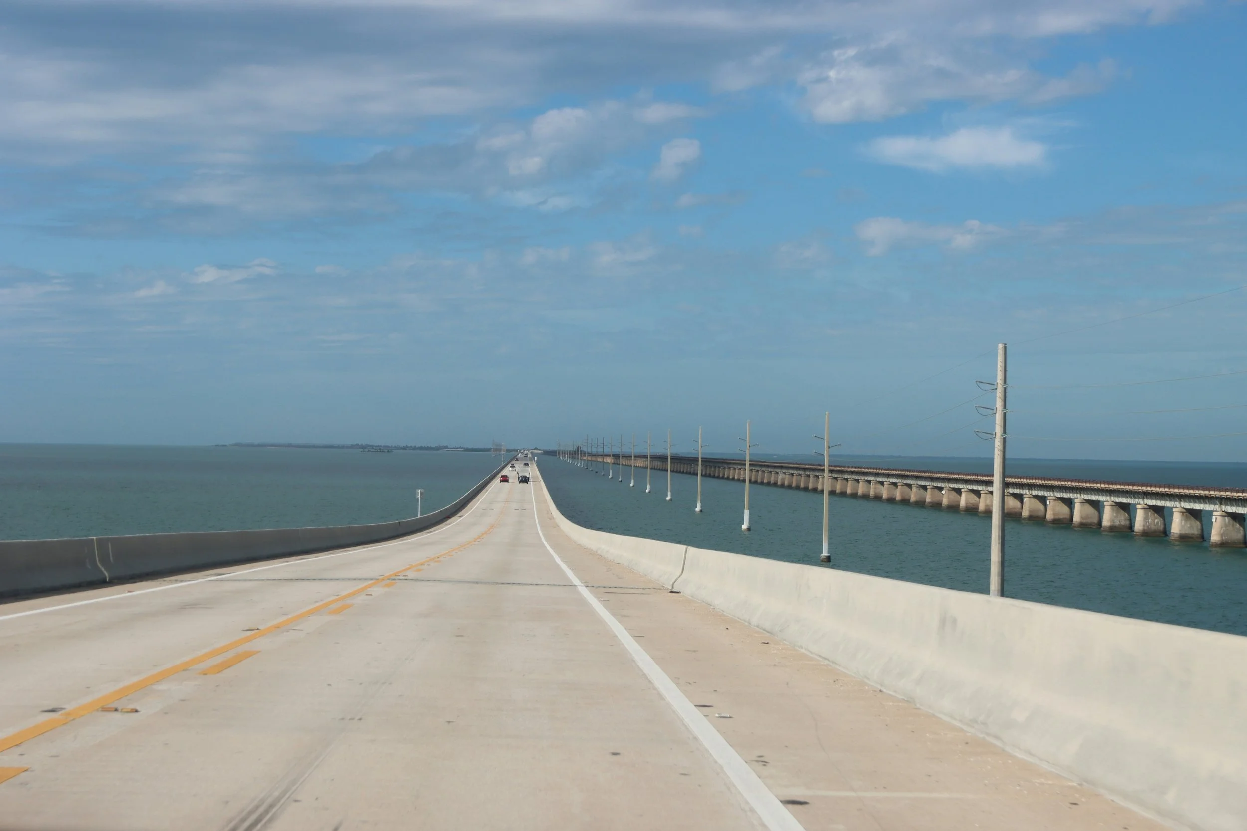 An image of a long bridge over water under a partly cloudy sky, with a few cars driving on it and utility poles running alongside.