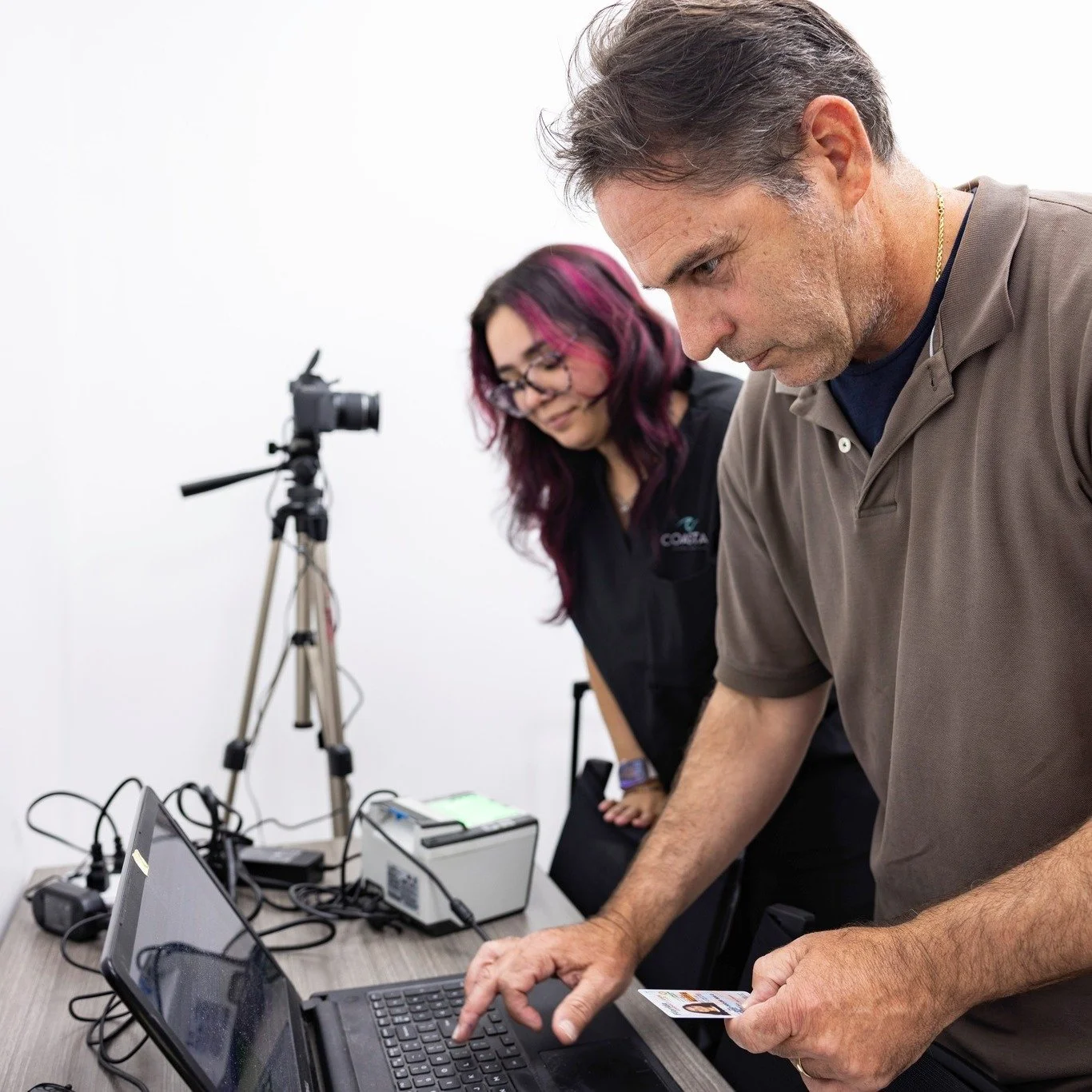 Two people working at a table with electronic equipment; a man is using a laptop, and a woman stands beside him holding a card, in a room with a camera on a tripod in the background.