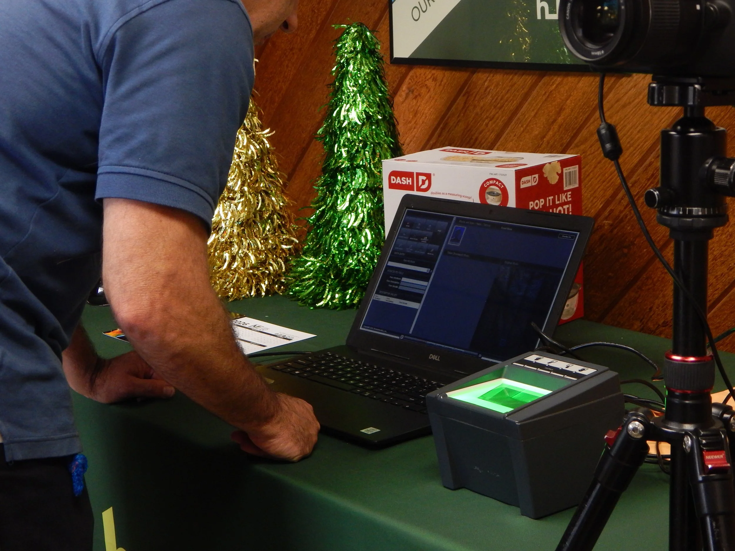 A person standing at a table with a laptop, a scanner, and festive holiday decorations, including three small decorated Christmas trees in gold and green, with a tree in the background and a box of Dash popcorn.