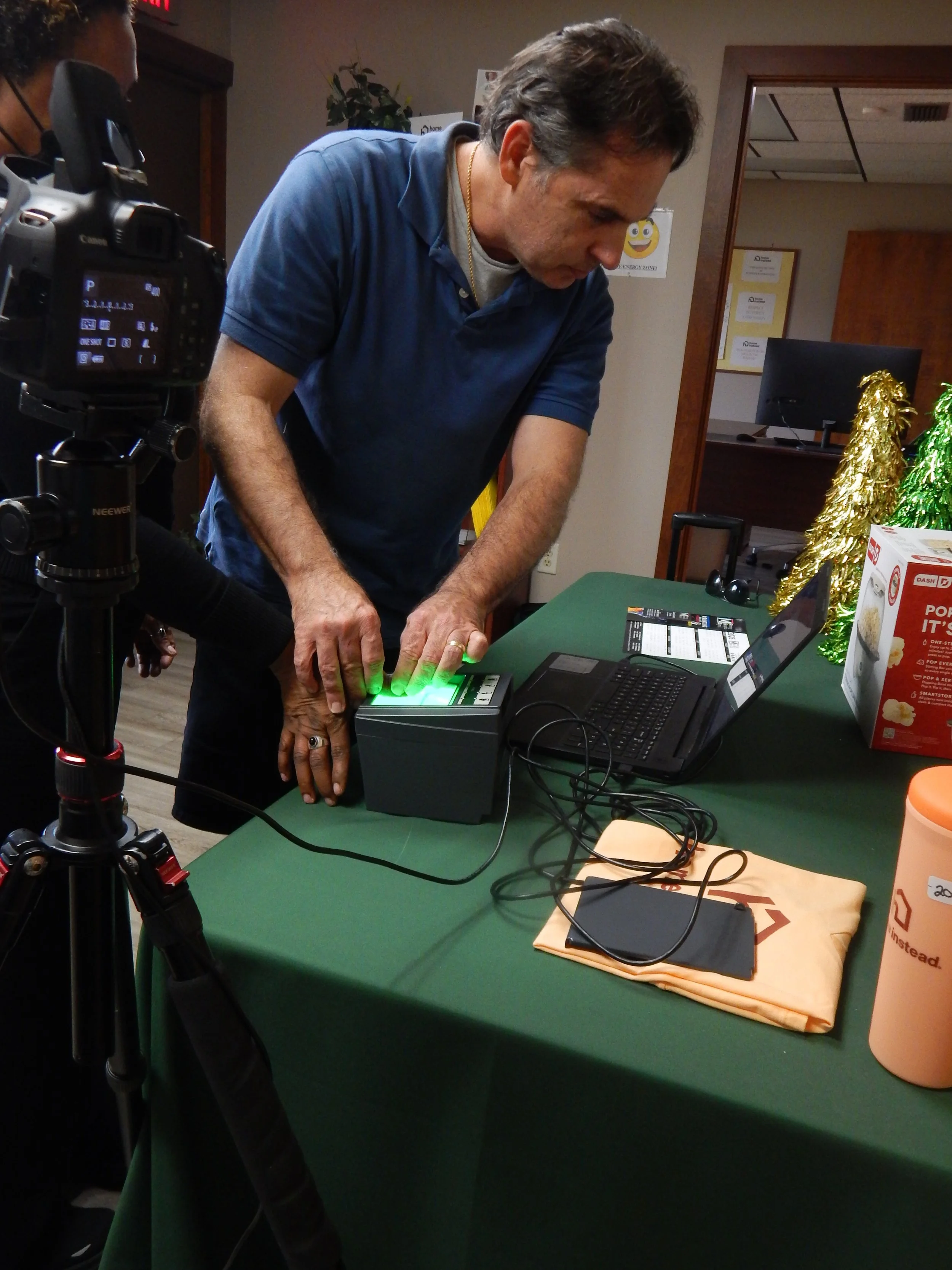 A man using a green sign-in device at a green-covered table with a laptop, smartphone, and holiday decorations nearby.