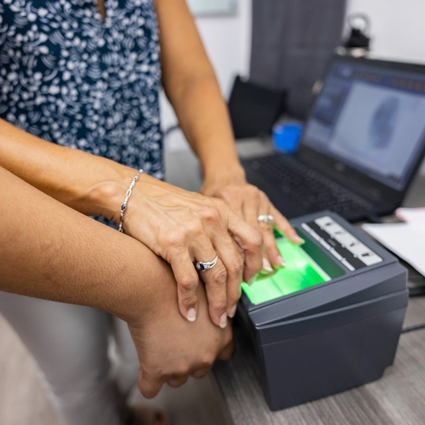Two women, one helping the other with a fingerprint scanner, with a laptop and office supplies in the background.