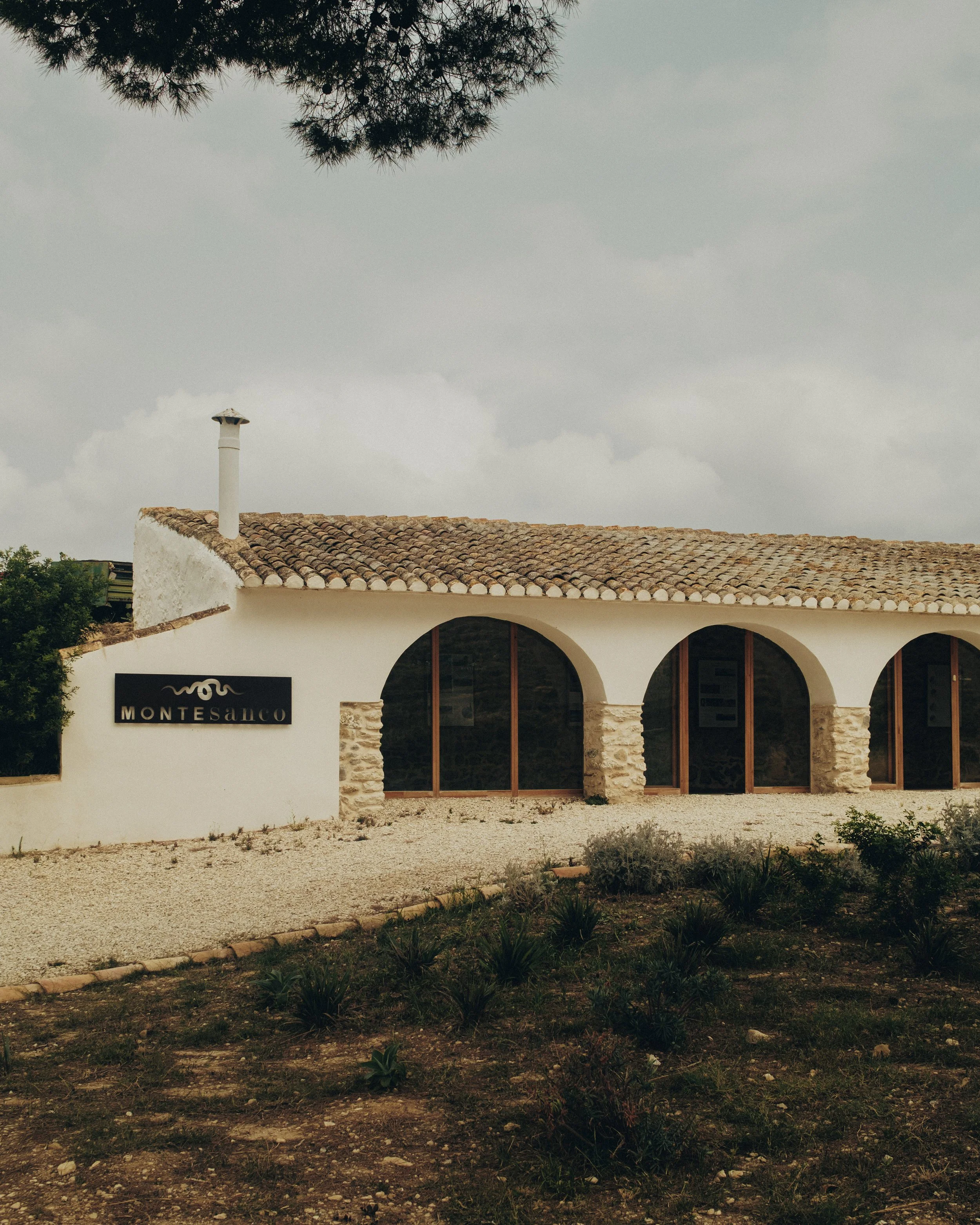 Front view of a white stucco building with a tiled roof, arched windows, and a sign reading 'Montesanco', with a gravel yard and bushes in the foreground under cloudy skies.
