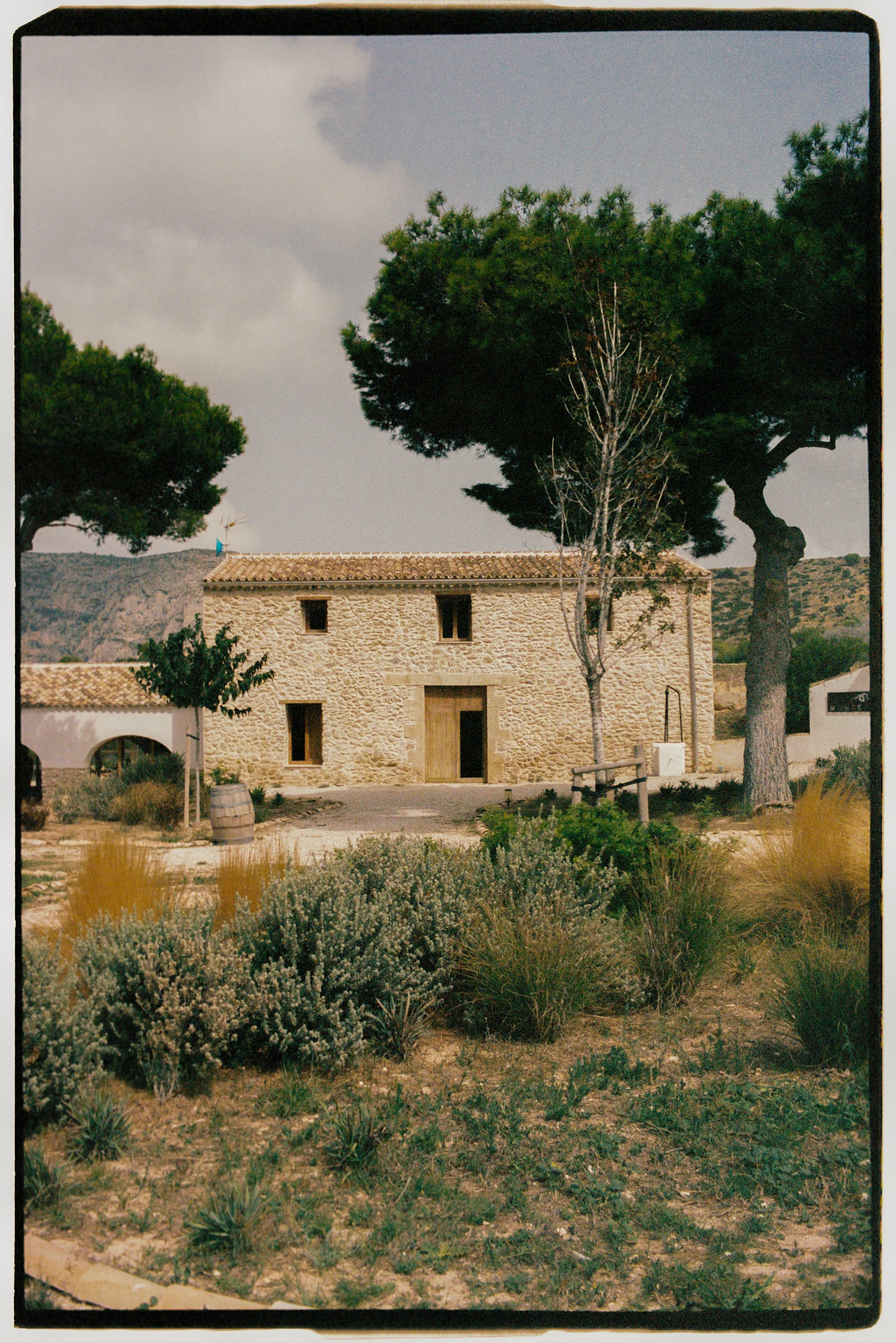 A stone house with a tiled roof and small windows, surrounded by desert-like plants and trees. The sky is partly cloudy, with hills in the background.