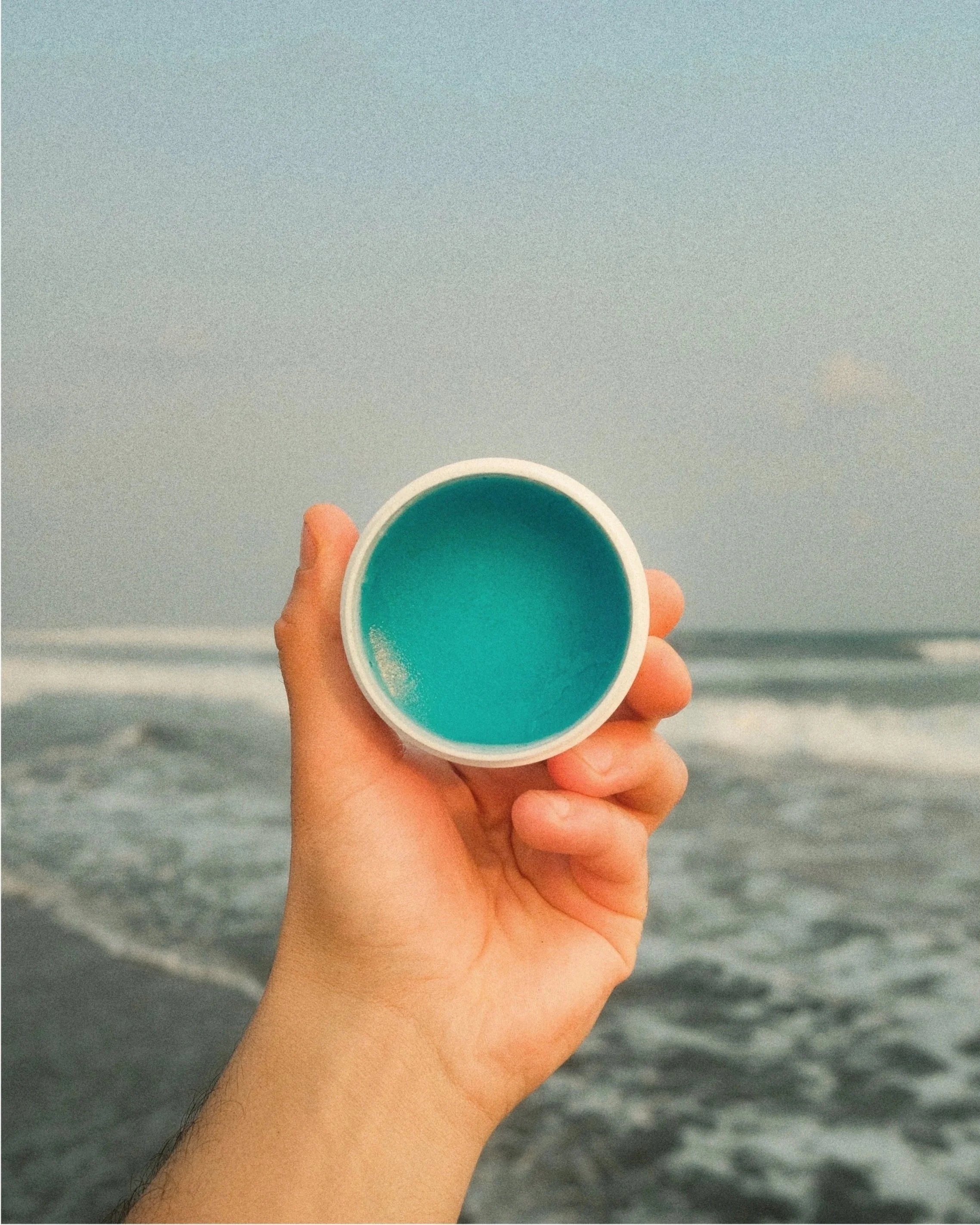 A person holding a white cup filled with blue liquid near the beach with the ocean in the background.