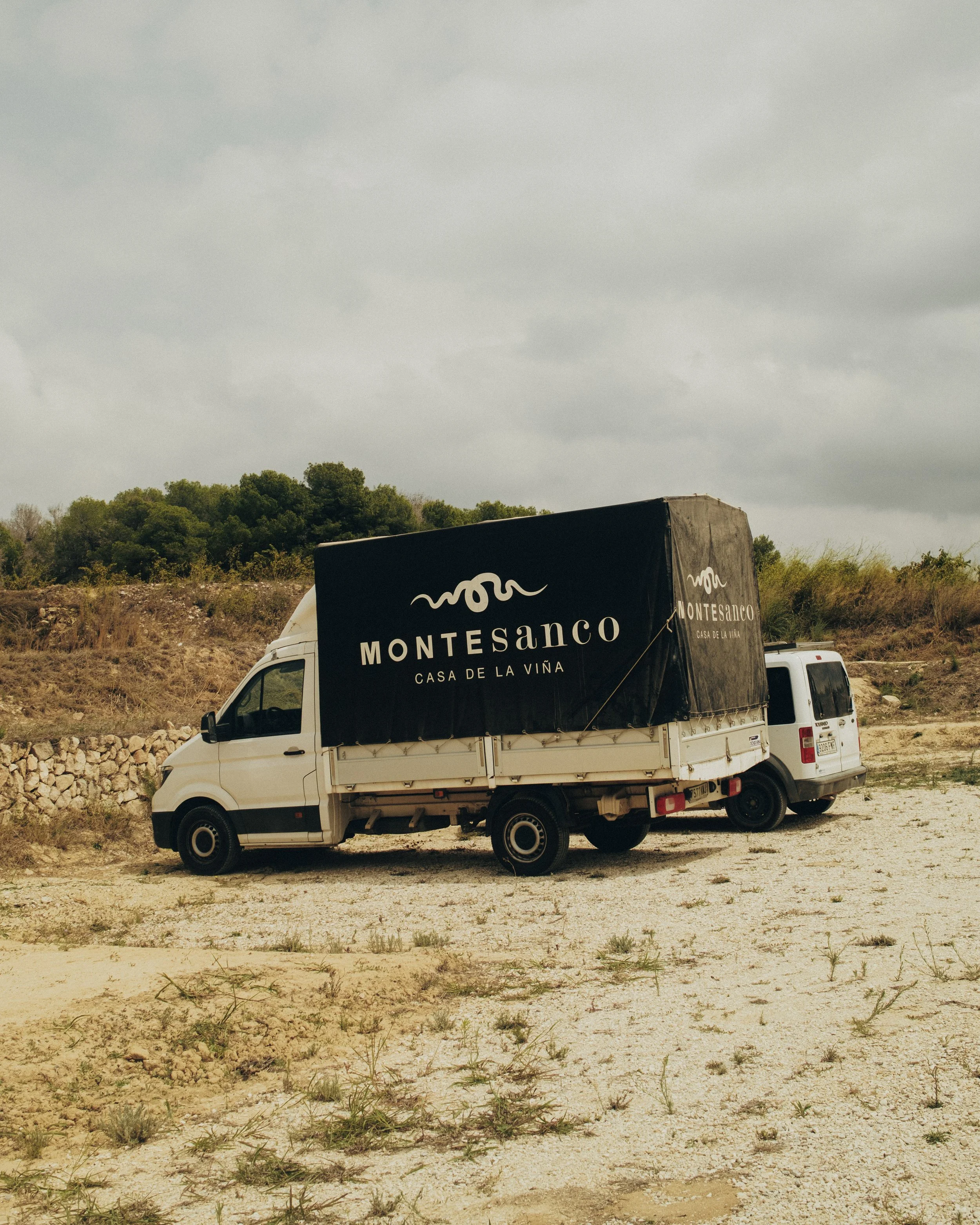 A white truck with a black trailer displaying the word "Montesanco" and the phrase "Casa de la Viña" parked on a dirt field with sparse grass, with trees and a cloudy sky in the background.