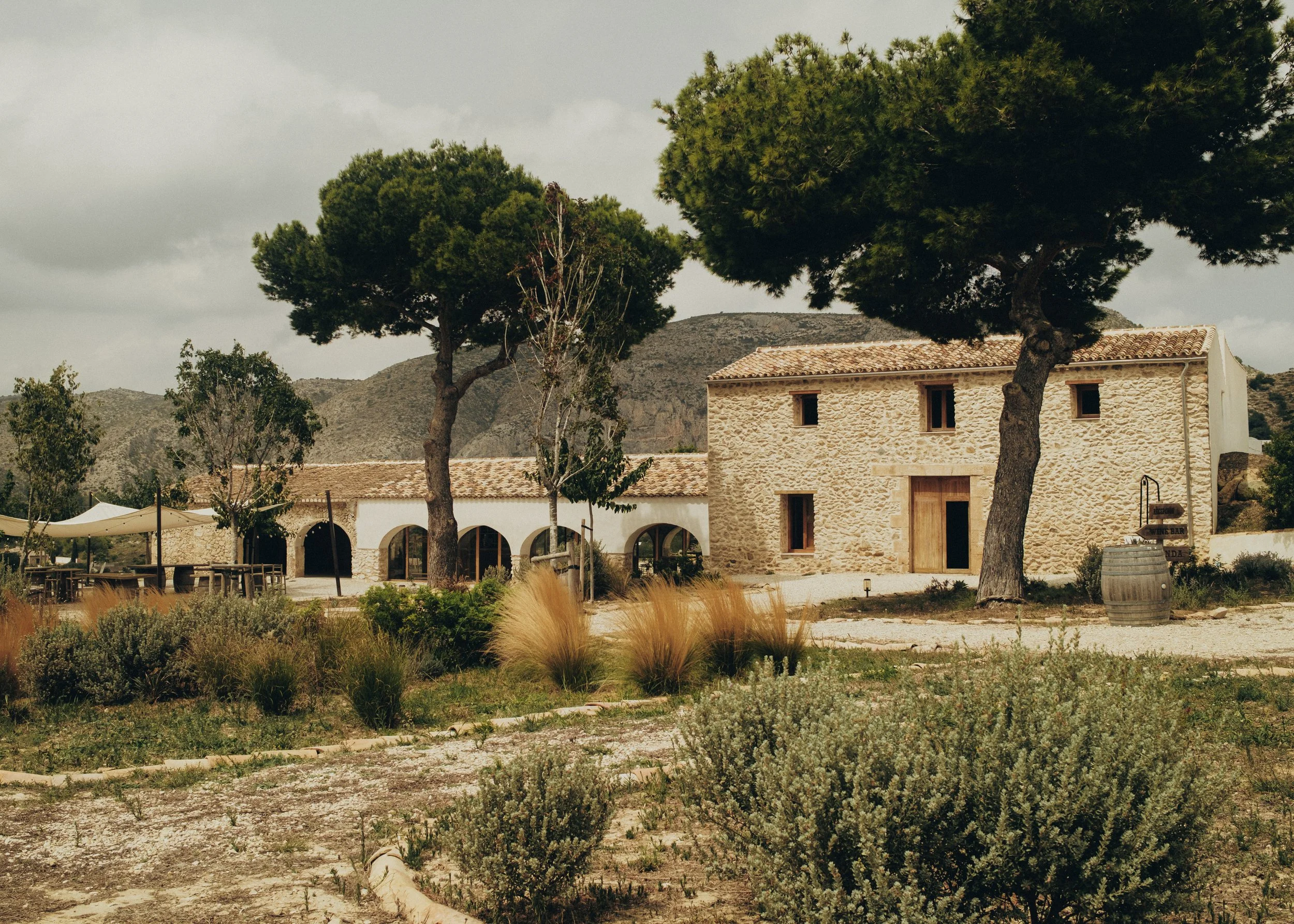 A stone house with a tiled roof surrounded by trees and bushes, with mountains in the background and an overcast sky.