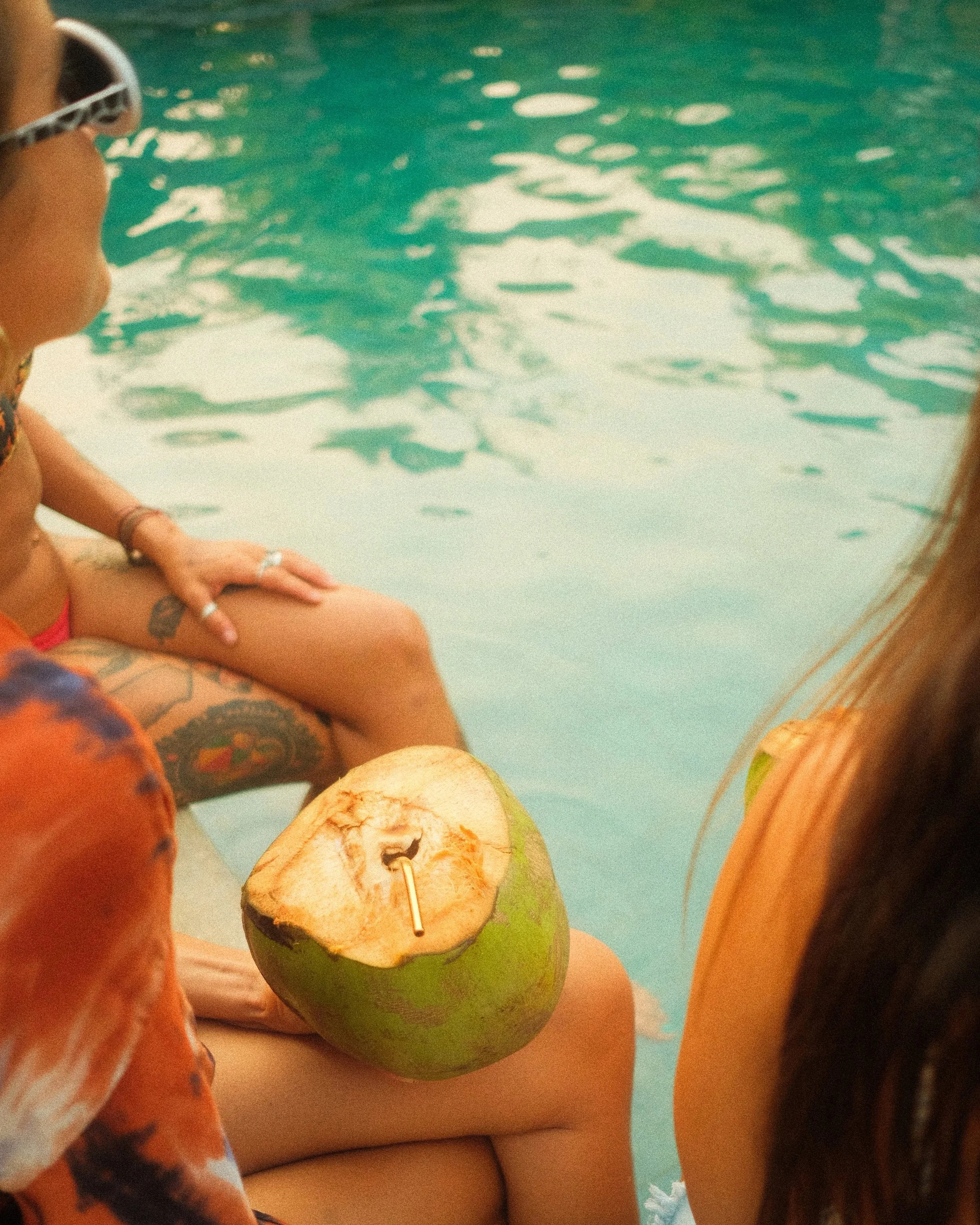 Person sitting by a pool, wearing sunglasses and a swimsuit, holding a coconut with a drinking straw, with another person nearby.