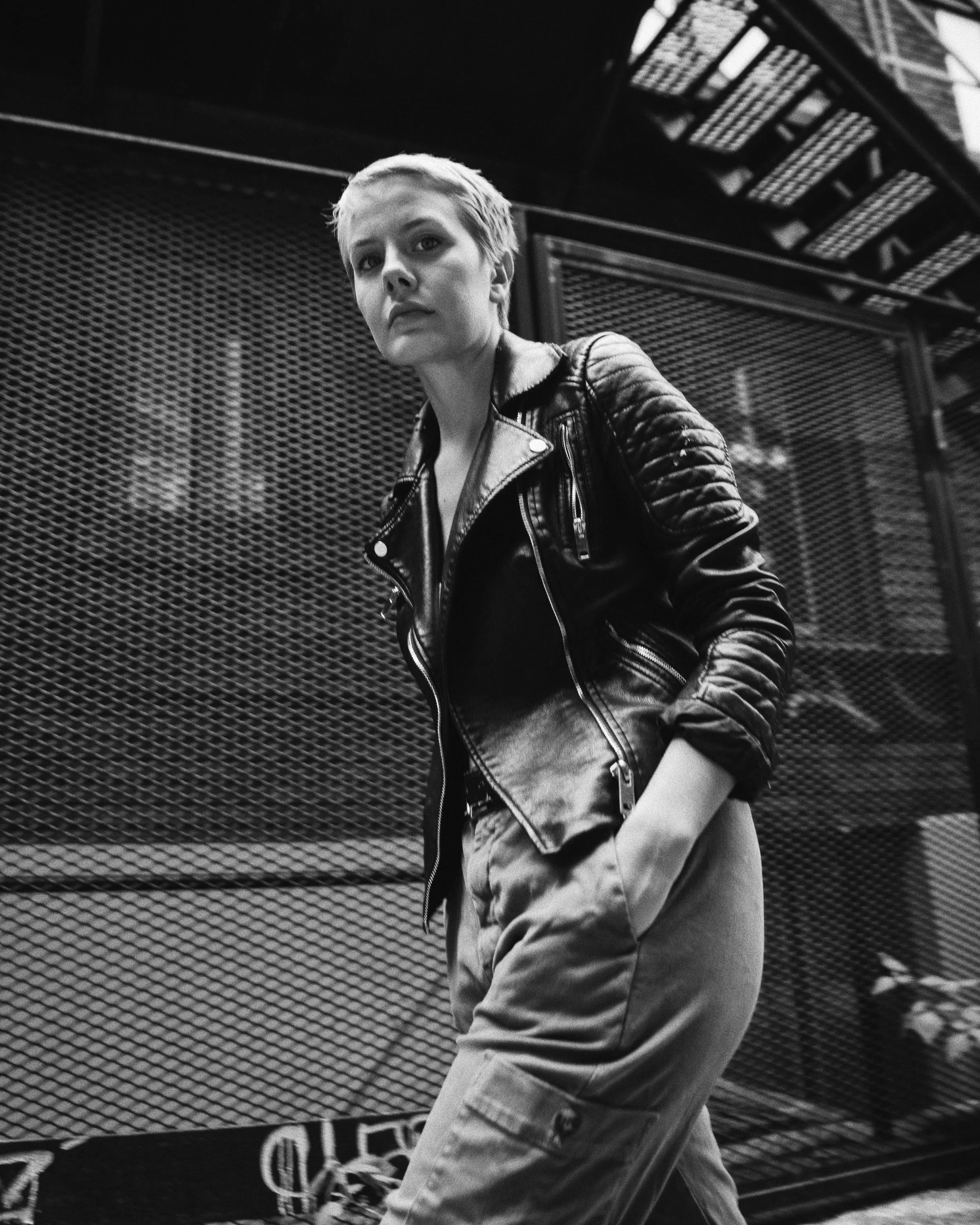 A woman with short blonde hair wearing a leather jacket and jeans standing in front of a chain-link fence.