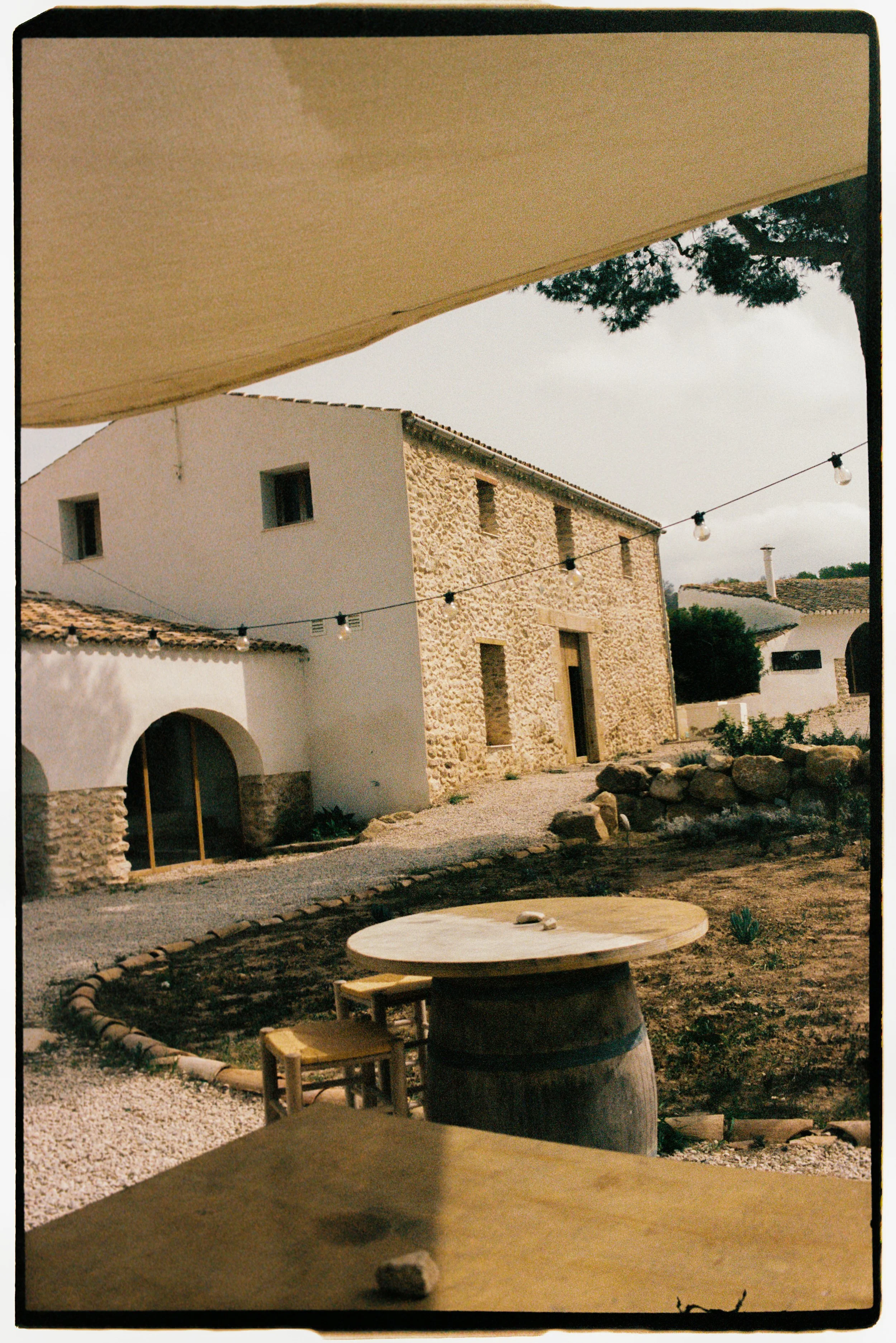 Outdoor scene with rustic stone and white buildings, string lights, a table made from a large barrel, and a pathway with gravel and rocks.