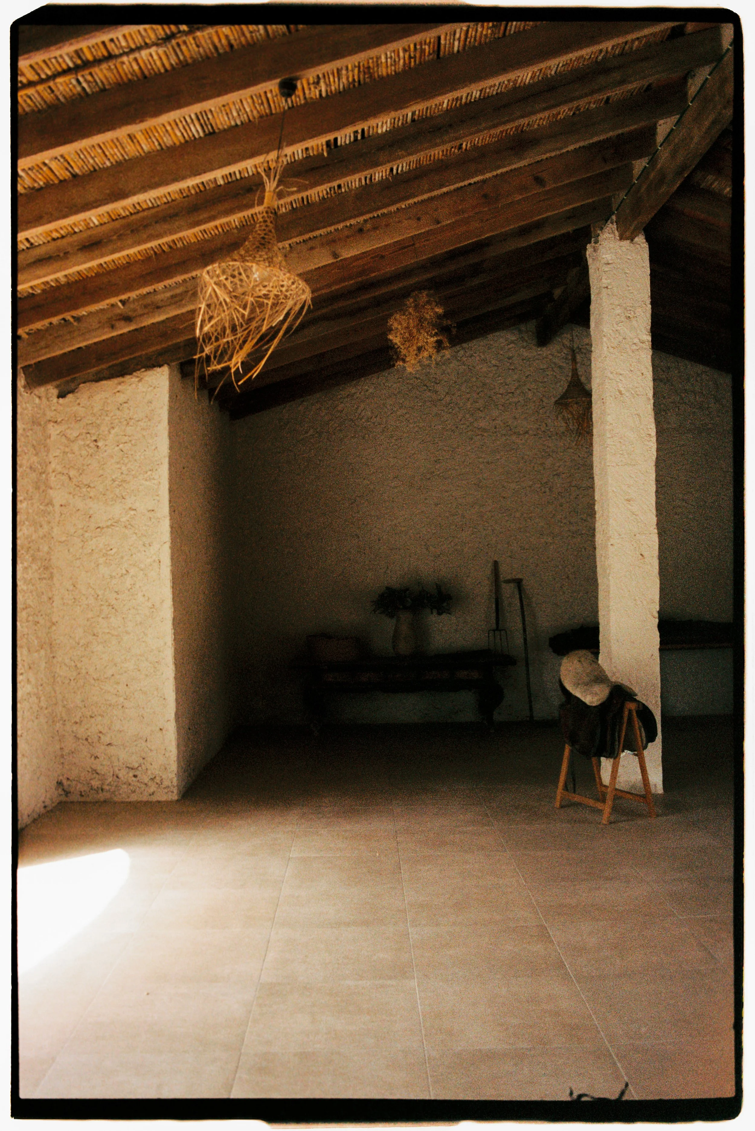 Interior of a rustic room with a wooden ceiling, stone wall, and tiled floor. A wicker pendant light hangs from the ceiling, and a small wooden table with decorative items is visible in the background. The room appears warmly lit, creating a cozy atmosphere.