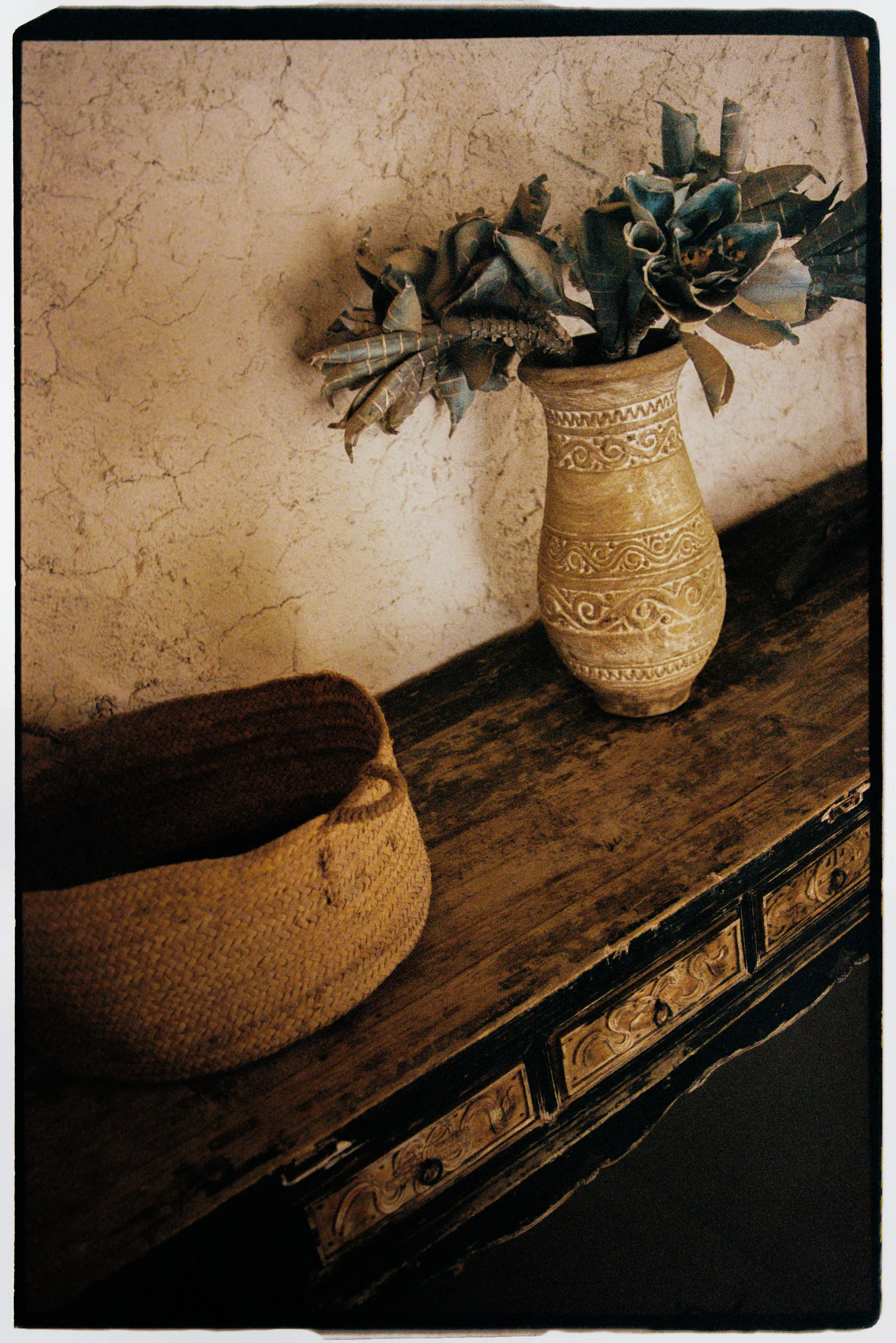 A tall, decorative ceramic vase filled with dried or artificial dark-colored leaves and flowers, placed on a wooden surface next to a woven basket, against a textured beige wall.
