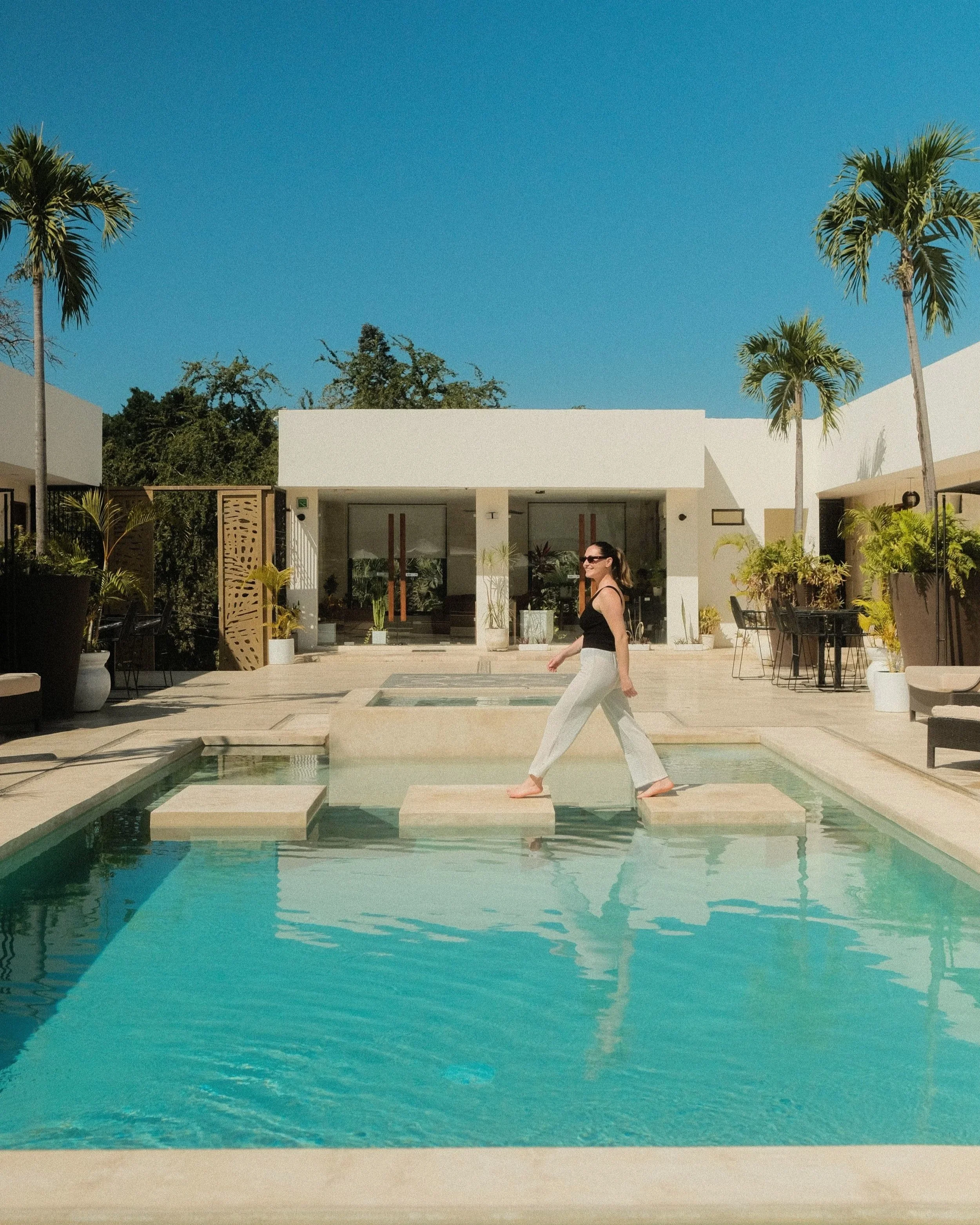 A woman in a black top and sunglasses walking near a swimming pool with a modern building and palm trees in the background on a sunny day.