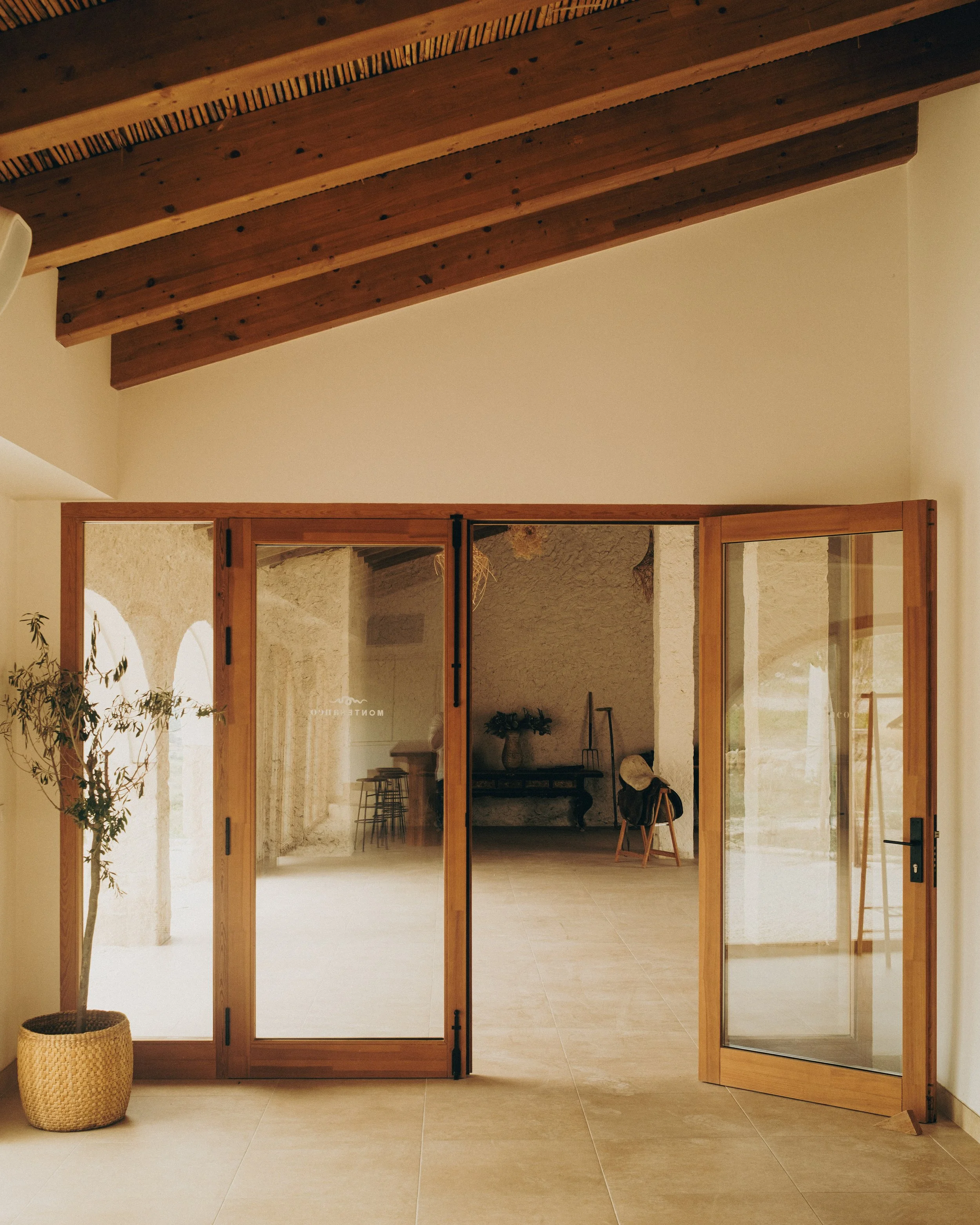 View of a room through wooden-framed glass sliding doors, with beige tiled floor, cream walls, and a slatted wooden ceiling. Inside, there is a table, two stools, and some decorative items, with natural light illuminating the space.