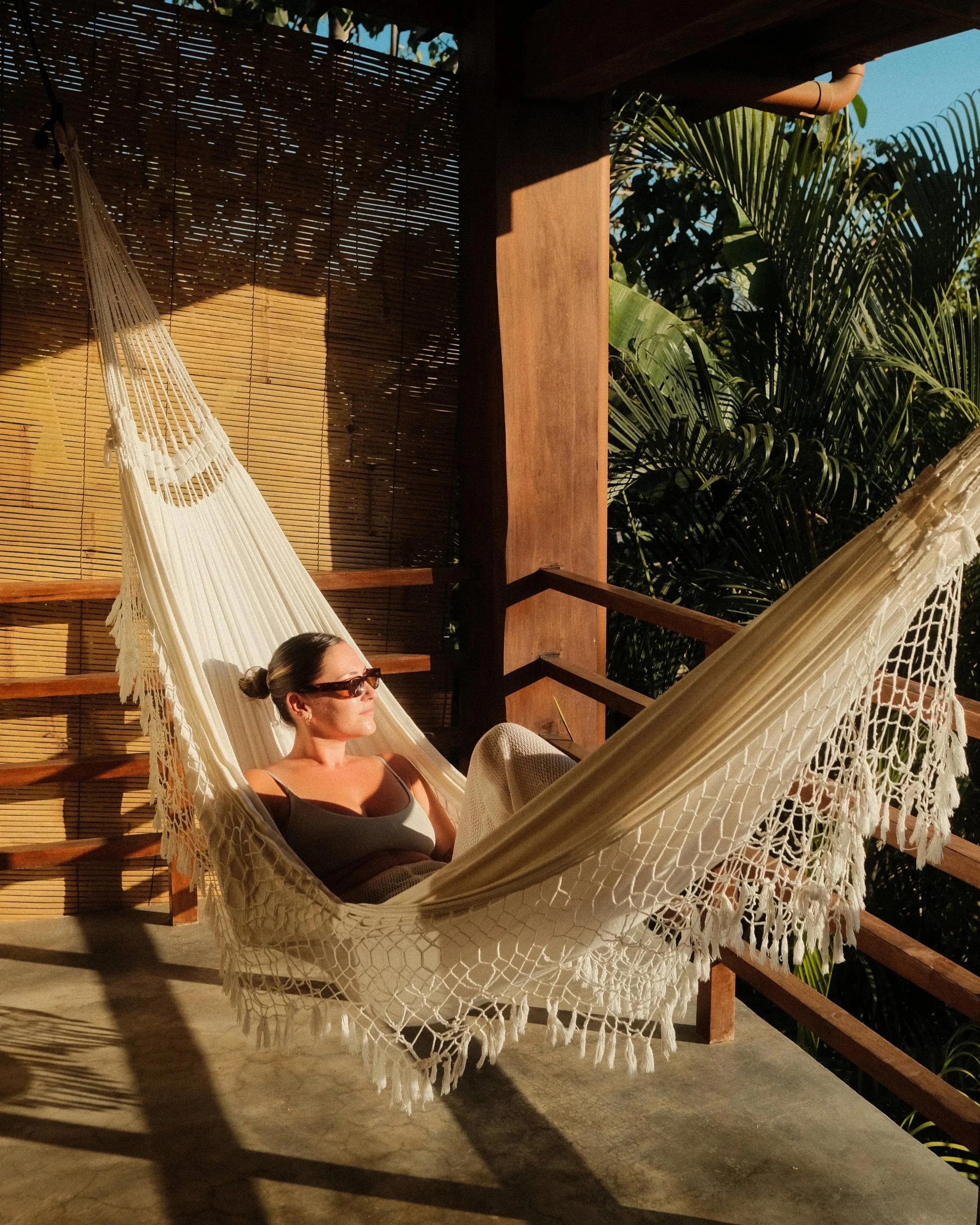 A woman relaxing in a white hammock outdoors with tropical plants in the background and sunlight casting shadows.