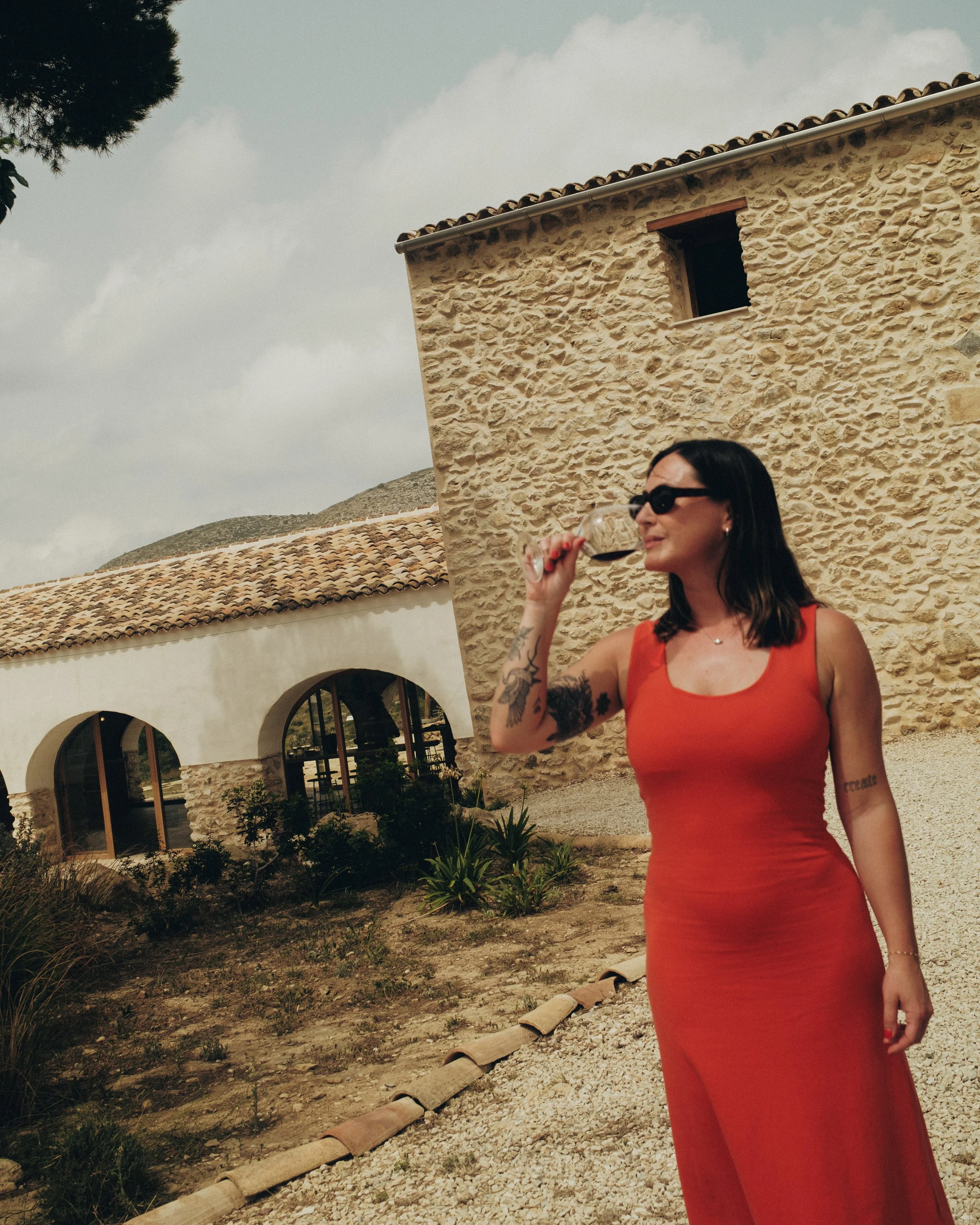 A woman with dark hair and tattoos on her arm, wearing sunglasses and an orange dress, drinks red wine outdoors in front of a rustic stone building with arched windows and a tiled roof.
