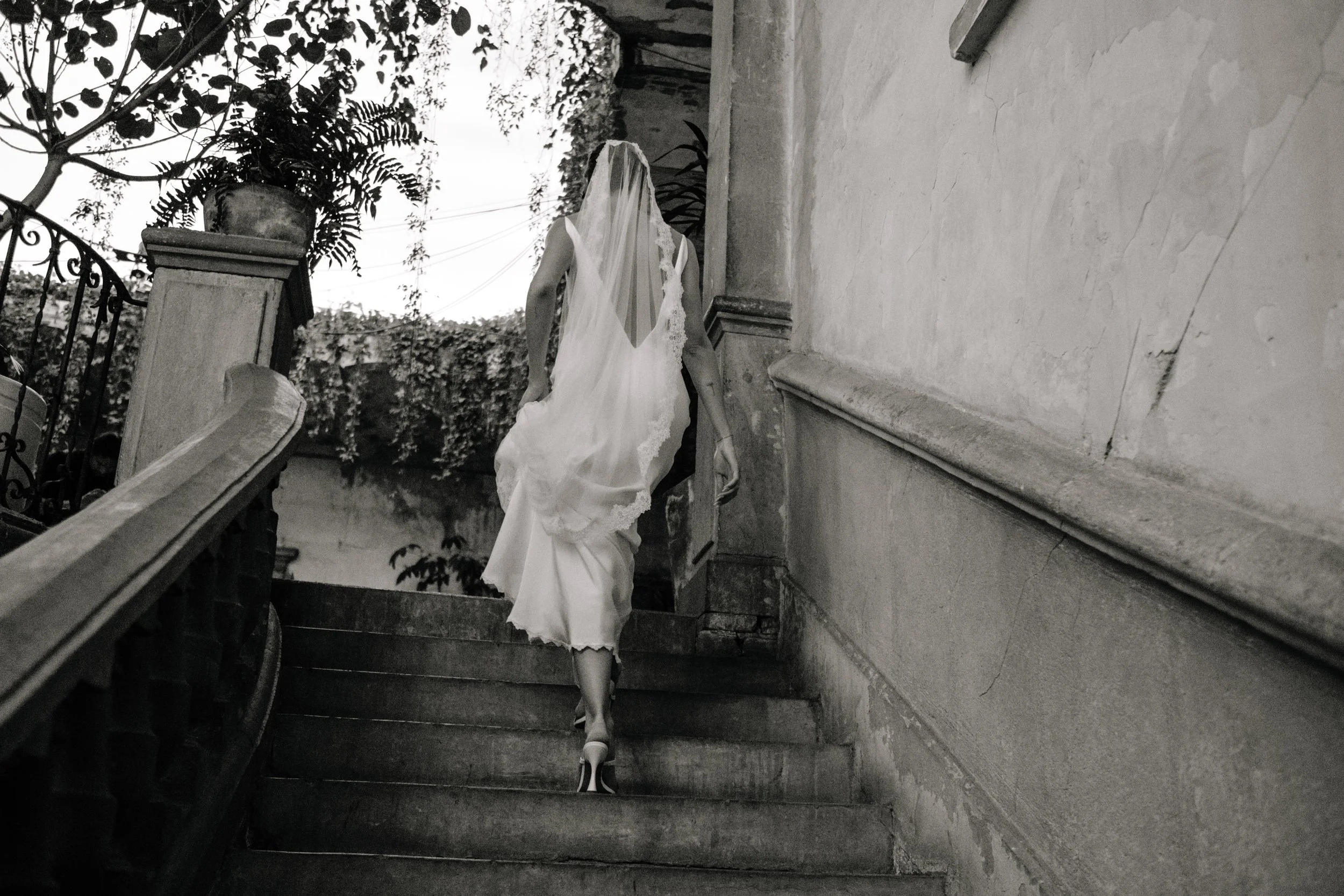 A woman in a wedding dress walks up an outdoor stone staircase with a rent veil covering her face and shoulders. The stairs are flanked by a stone wall and iron railing, and trees and foliage are visible in the background.