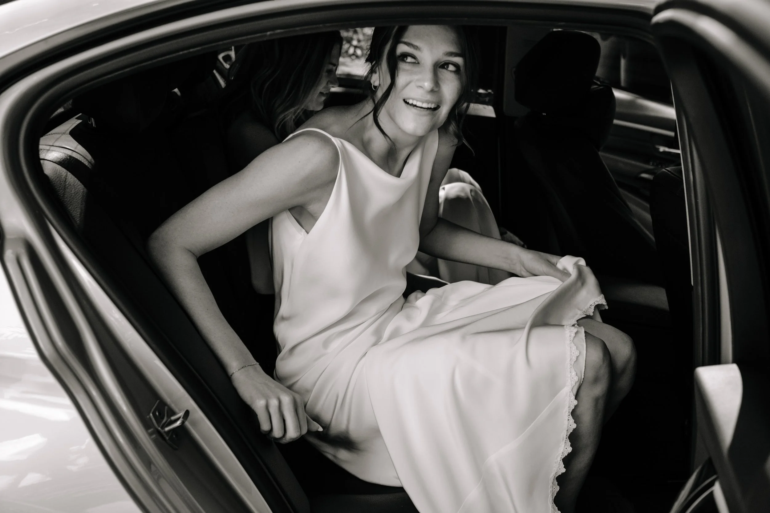 A woman in a wedding dress is getting into a car, smiling and looking out.