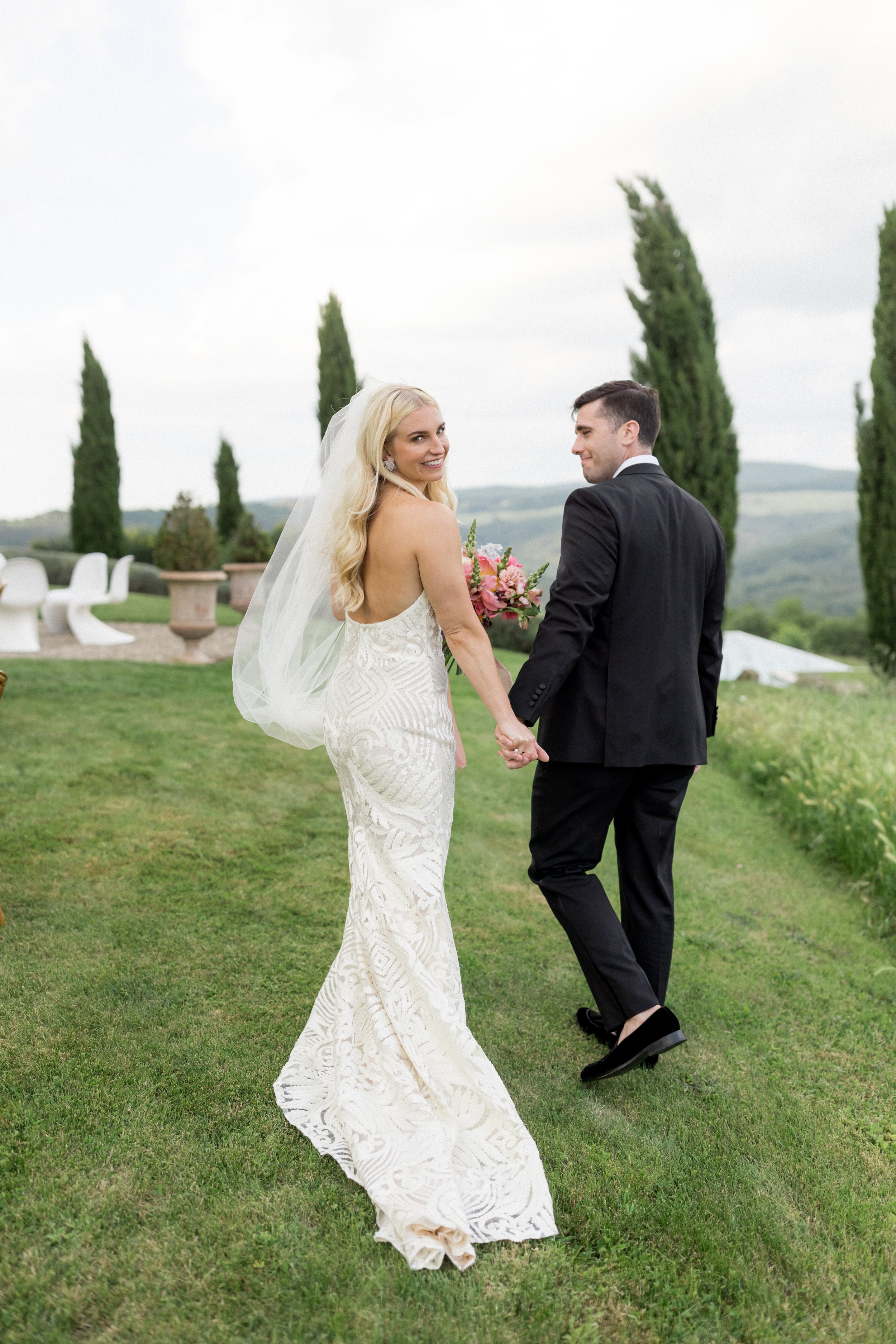 Bride and groom holding hands and walking in a garden on their wedding day, with scenic hills and cypress trees in the background.