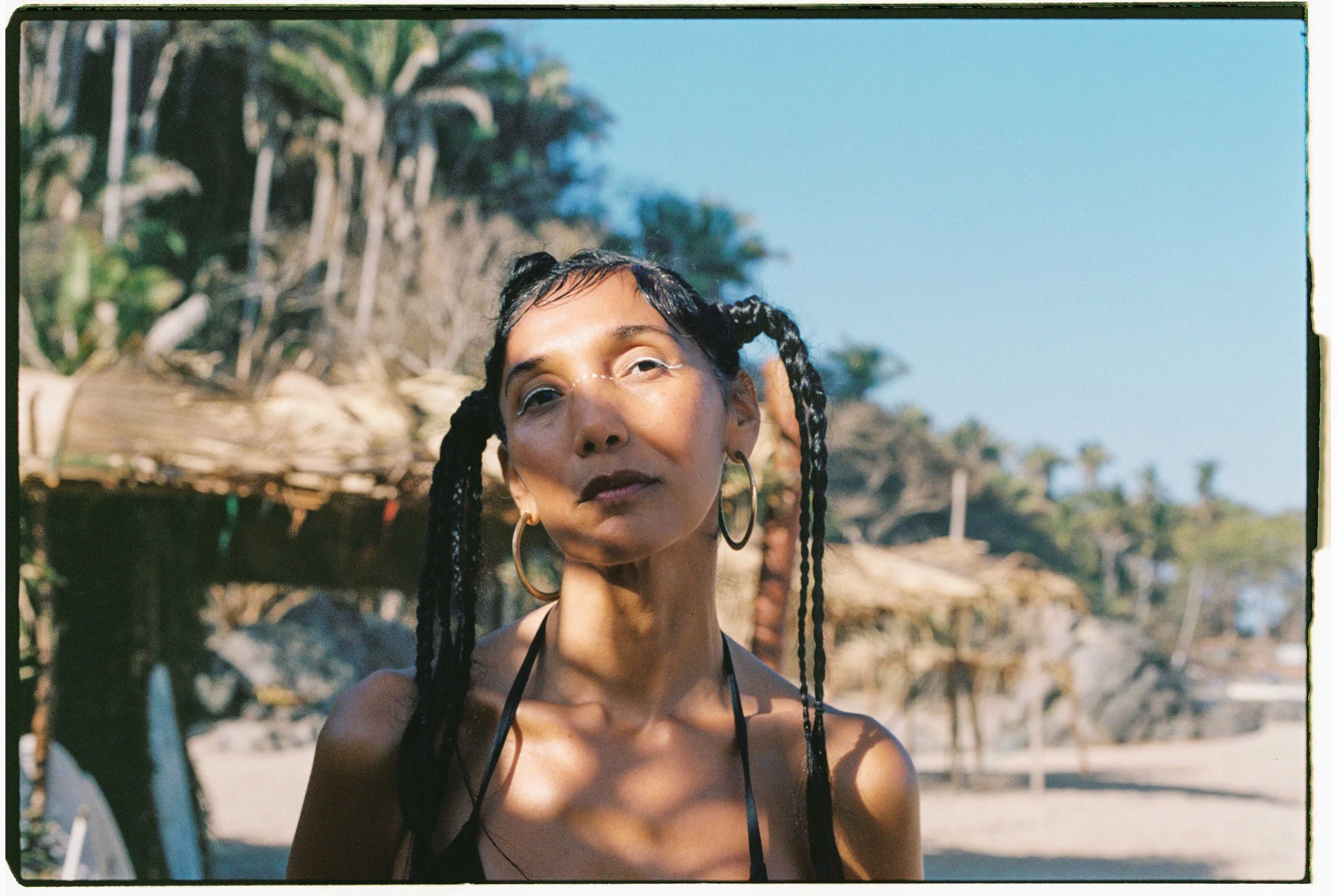 Woman with braided hair and hoop earrings standing outdoors with trees and beach huts in the background.